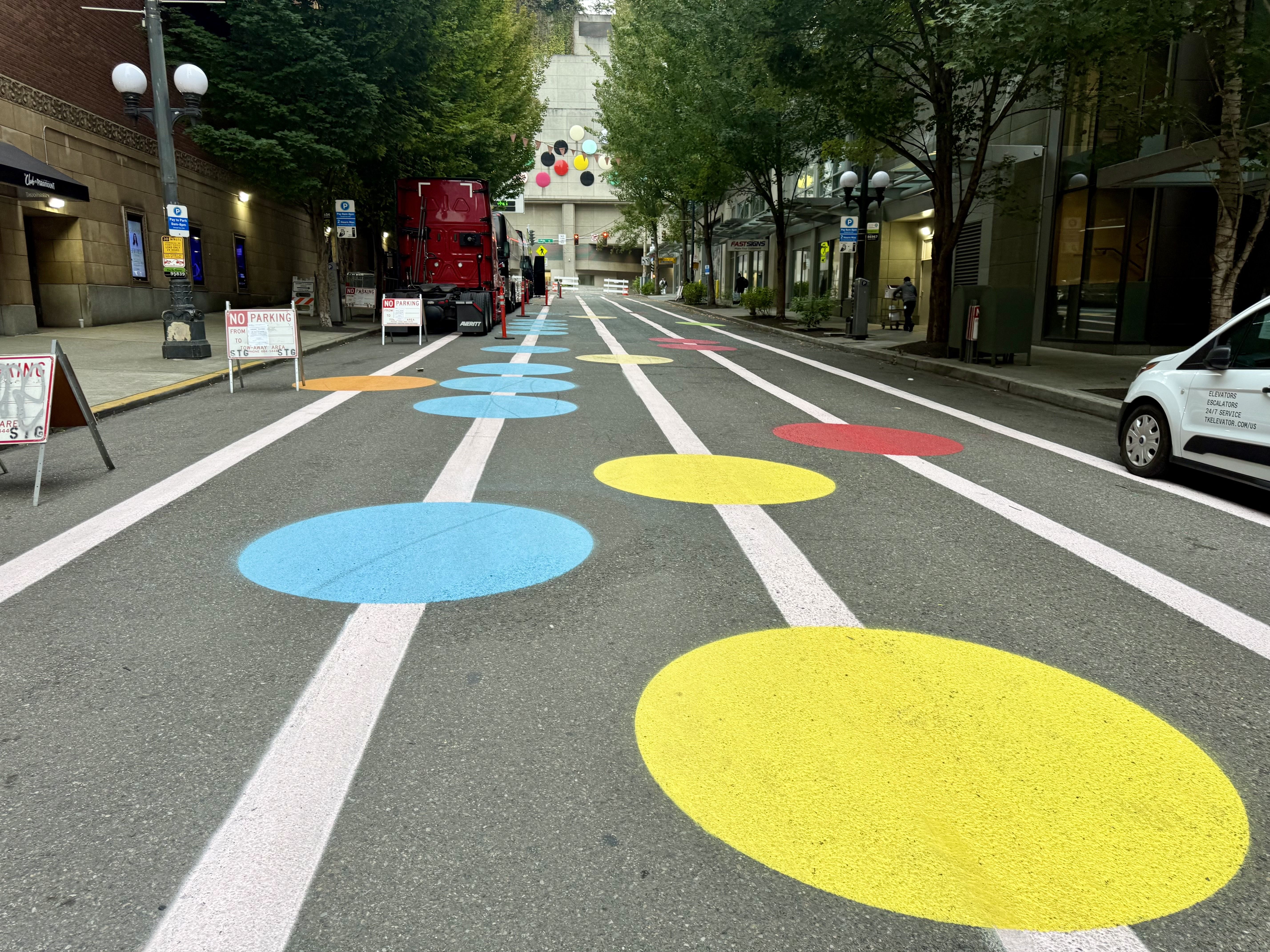Wide city street with trees and buildings. A red truck on the left; the road shows large blue, yellow, and red circular marks between white lane lines, with signs and people nearby.