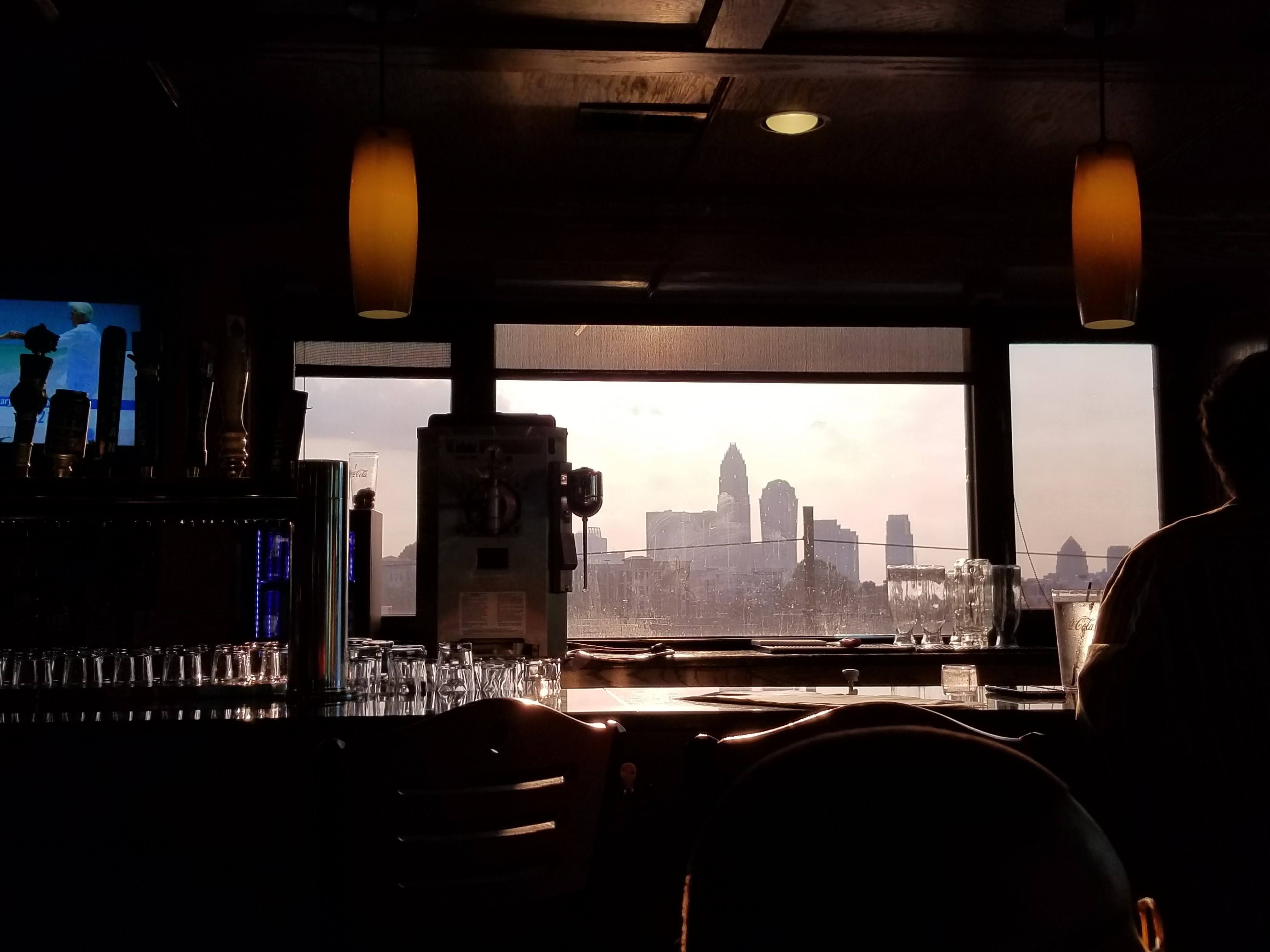 Dimly lit bar interior at sunset with glowing pendant lights, glasses on the counter, a TV on the left, and a city skyline visible through the large window.