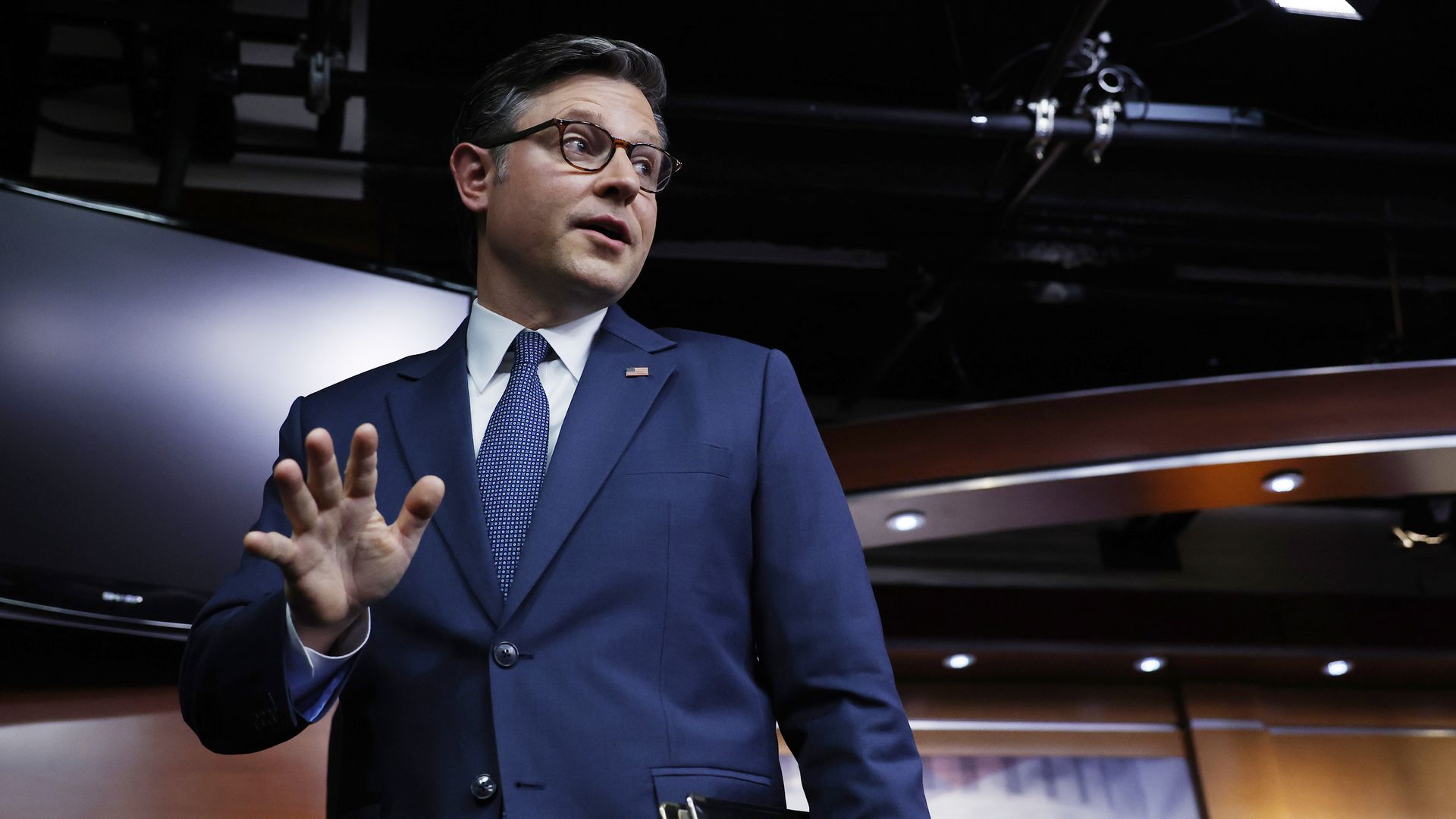 House Speaker Mike Johnson, wearing a blue suit and standing in a wooden TV gallery.
