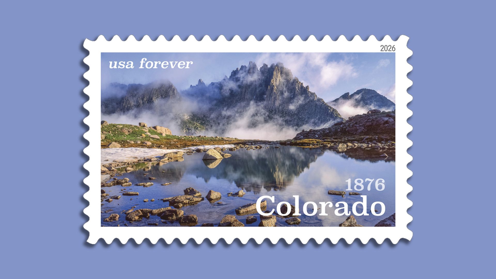 USA Forever Colorado stamp showing a rocky mountain peak with clouds and its reflection in a calm lake, surrounded by rocks and greenery, under a blue sky.