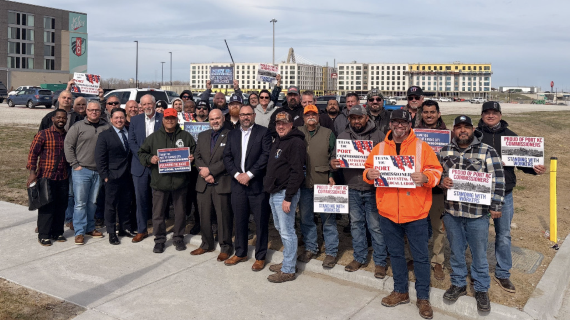 Group of diverse people posing outdoors on a sidewalk, holding signs in support of Port KC commissioners, with a construction site and new buildings behind them.