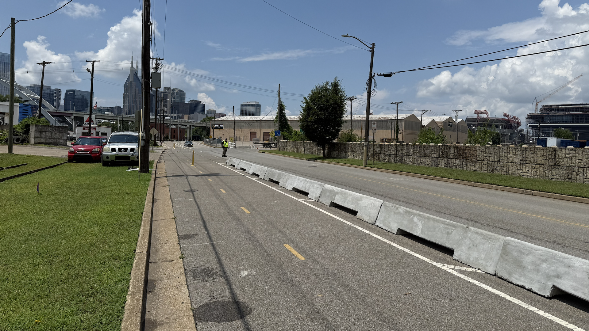Wide city street with utility poles and wires, a worker in high-vis near median barriers, construction site on the right, skyline buildings in distance under a blue sky.
