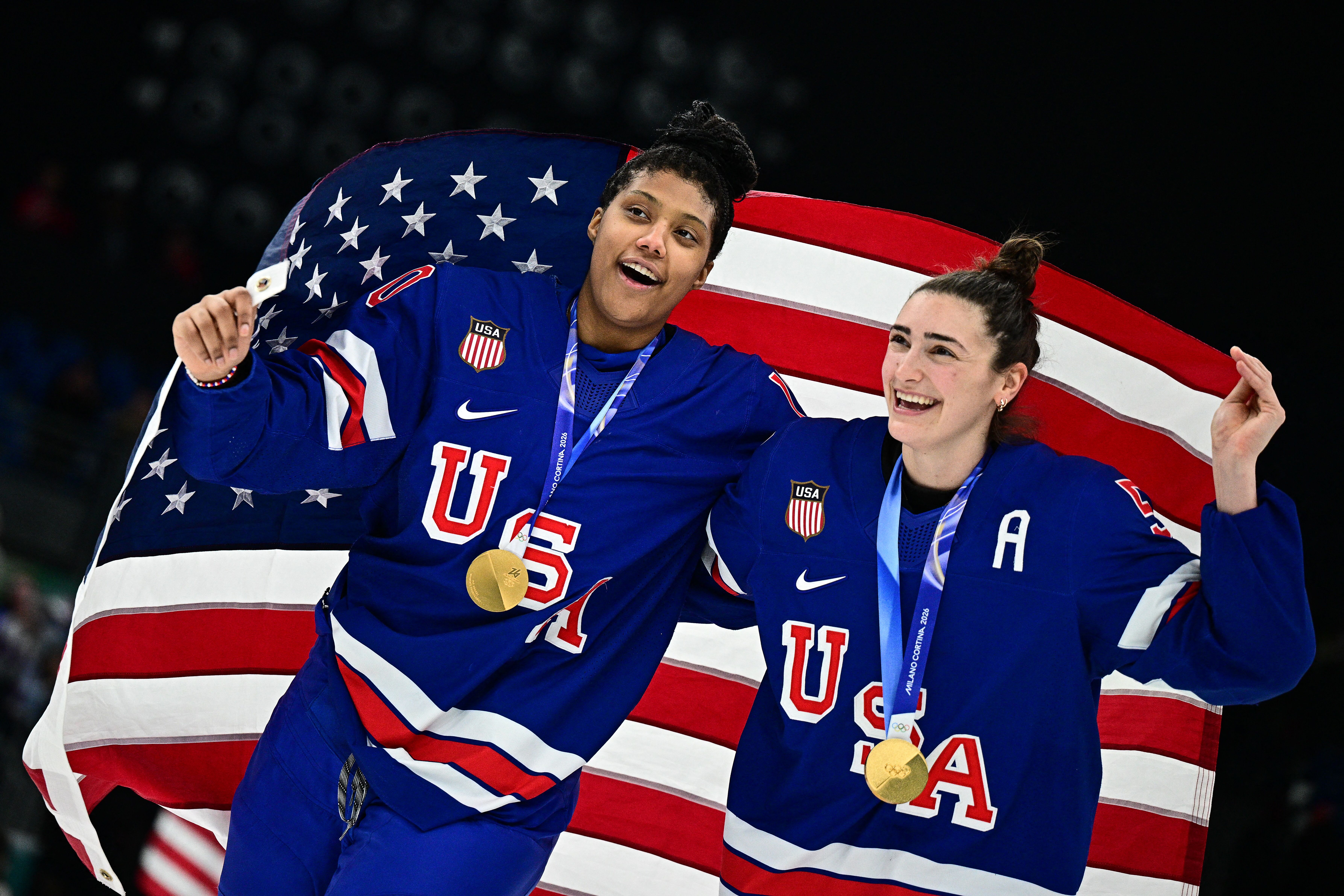 Team USA hockey players Laila Edwards and Megan Keller in blue jerseys with gold medals, smiling and holding an American flag behind them in a victory celebration.