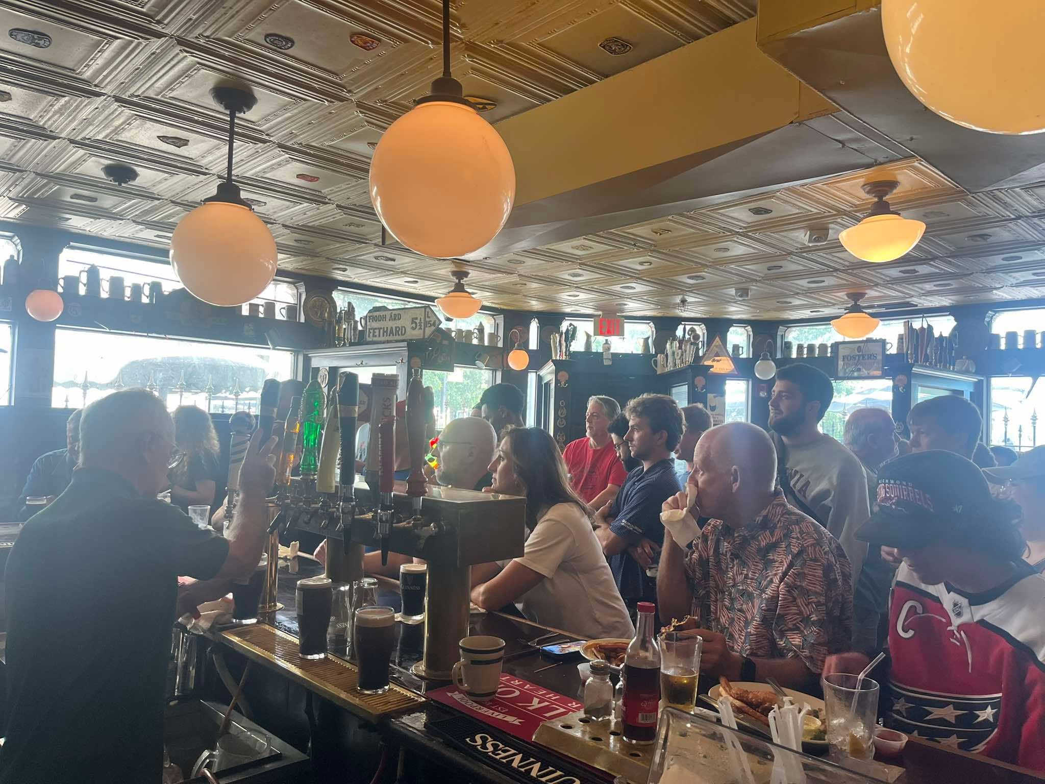 A crowded bar scene with people sitting at a long counter, beer taps in the foreground, and warm orange globe lights hanging from a tin ceiling. Windows line the back wall.