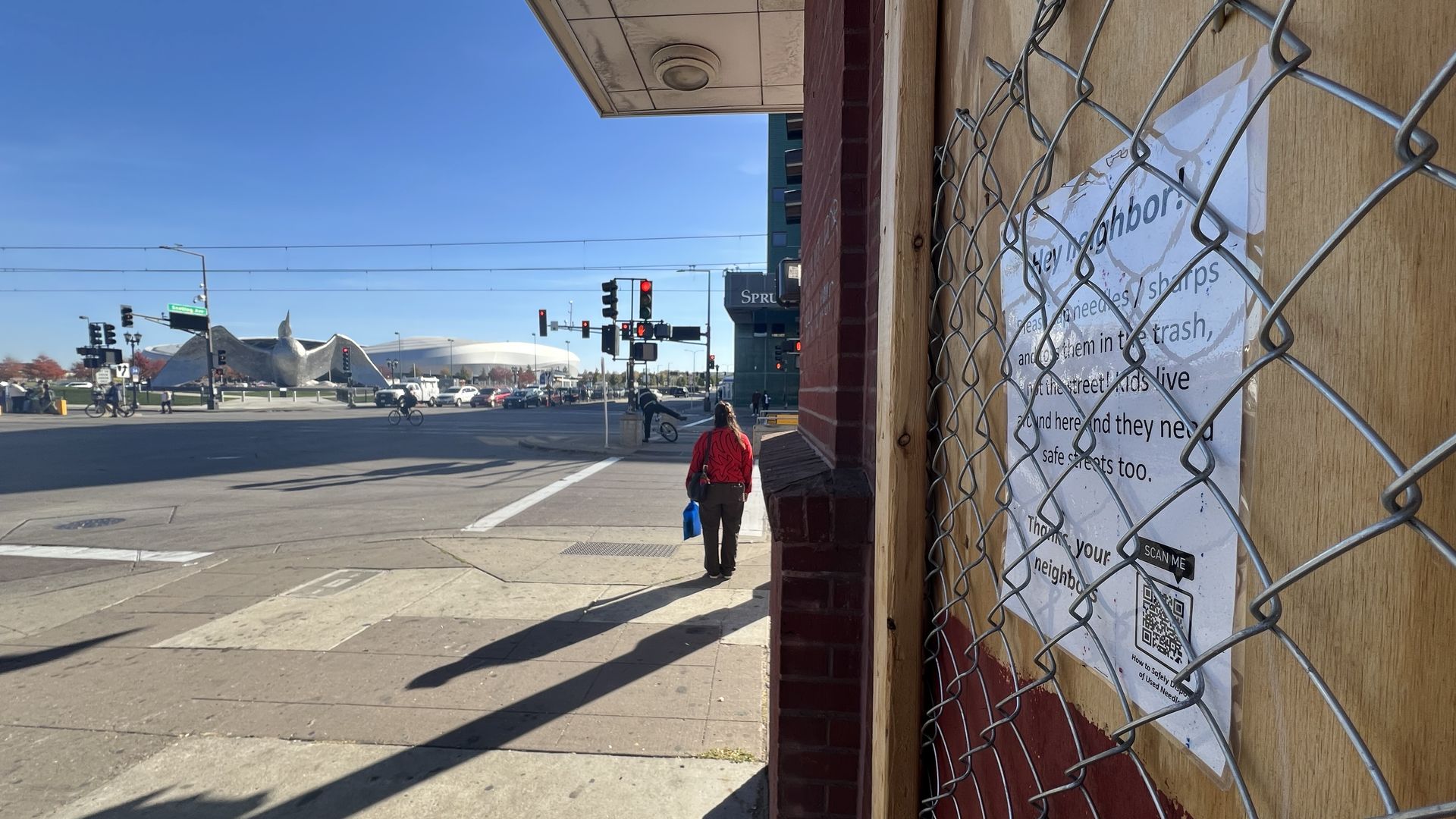 A boarded up window with a chain-link fence and a sign reading "Hey neighbor! Please cap needles/sharps and toss them in the trash, not the street! Kids live around here and they need safe streets too." A soccer stadium and loon sculpture stand in the background.