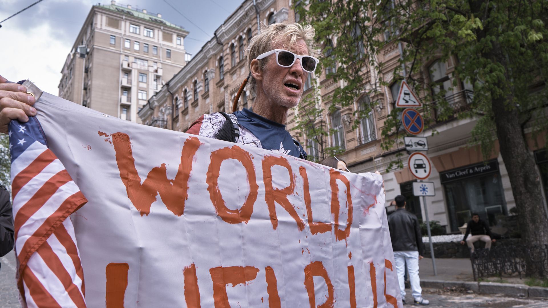 A man wearing white sunglasses carries a white banner with the words "WORLD HELP US" and an American flag.