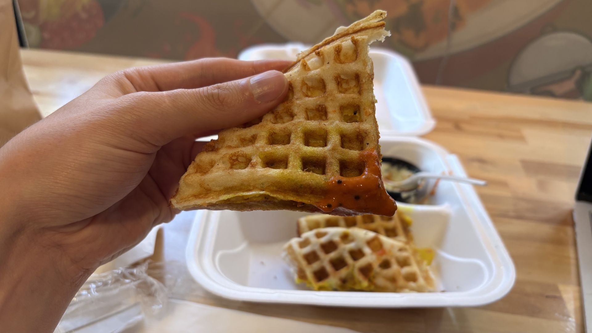 Hand holding a triangular piece of golden-brown waffle with red sauce on the edge, with a white takeout container holding more waffle pieces on a wooden table.