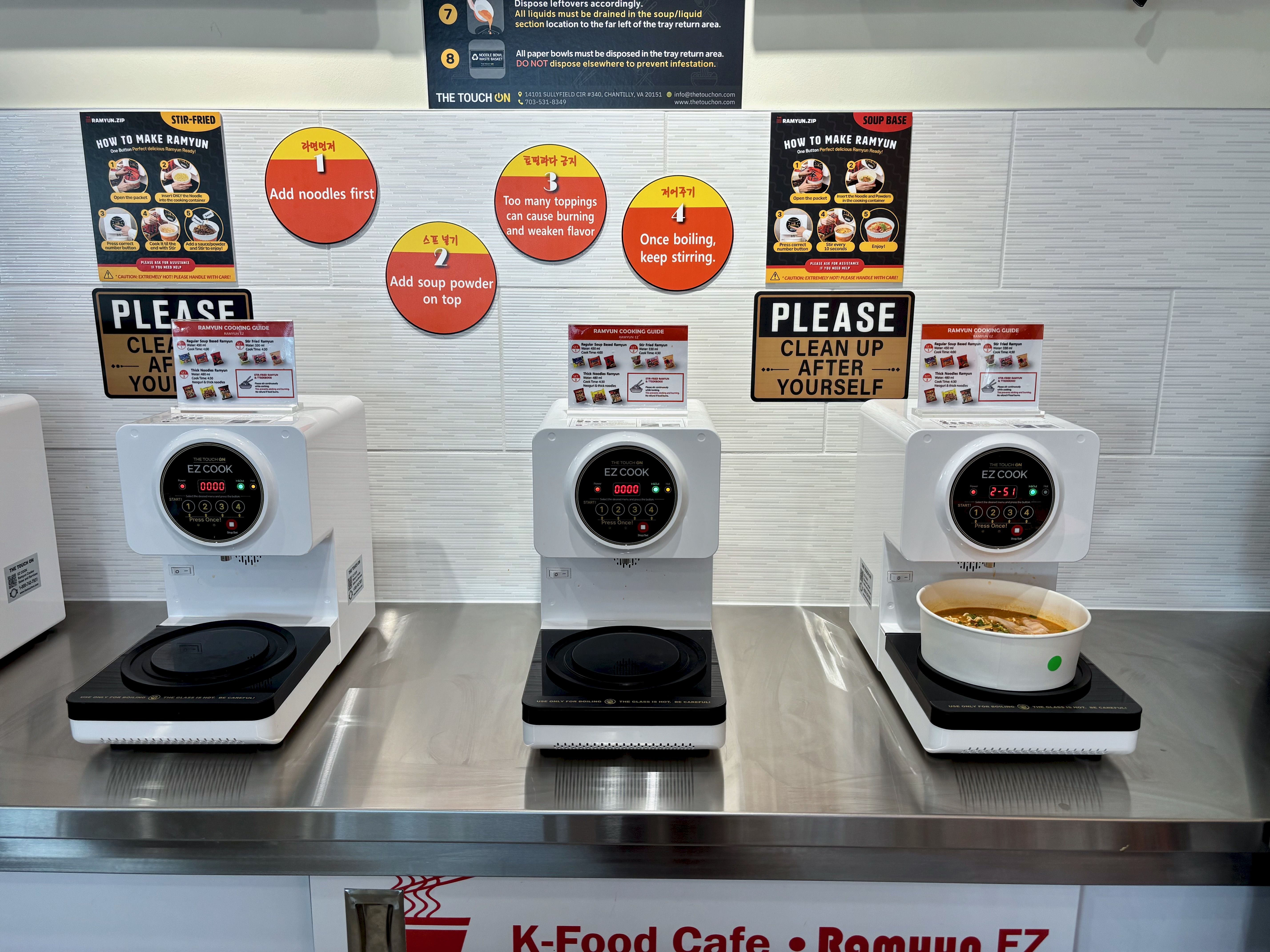 Row of white ramen cookers with digital displays on a counter, posters showing how to make ramen on tiled wall, and a big PLEASE CLEAN UP AFTER YOURSELF sign; a bowl of ramen on the right.