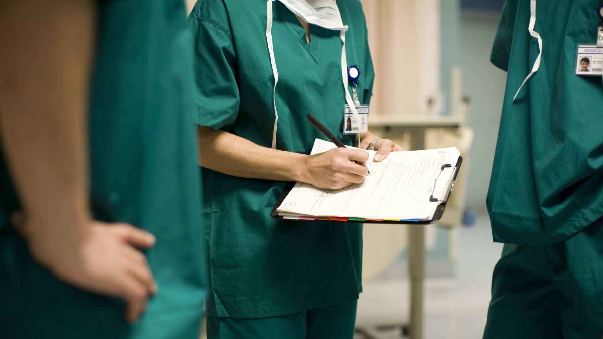 In this image, three doctors stand in green scrubs in a hallway. One is holding a clipboard. 