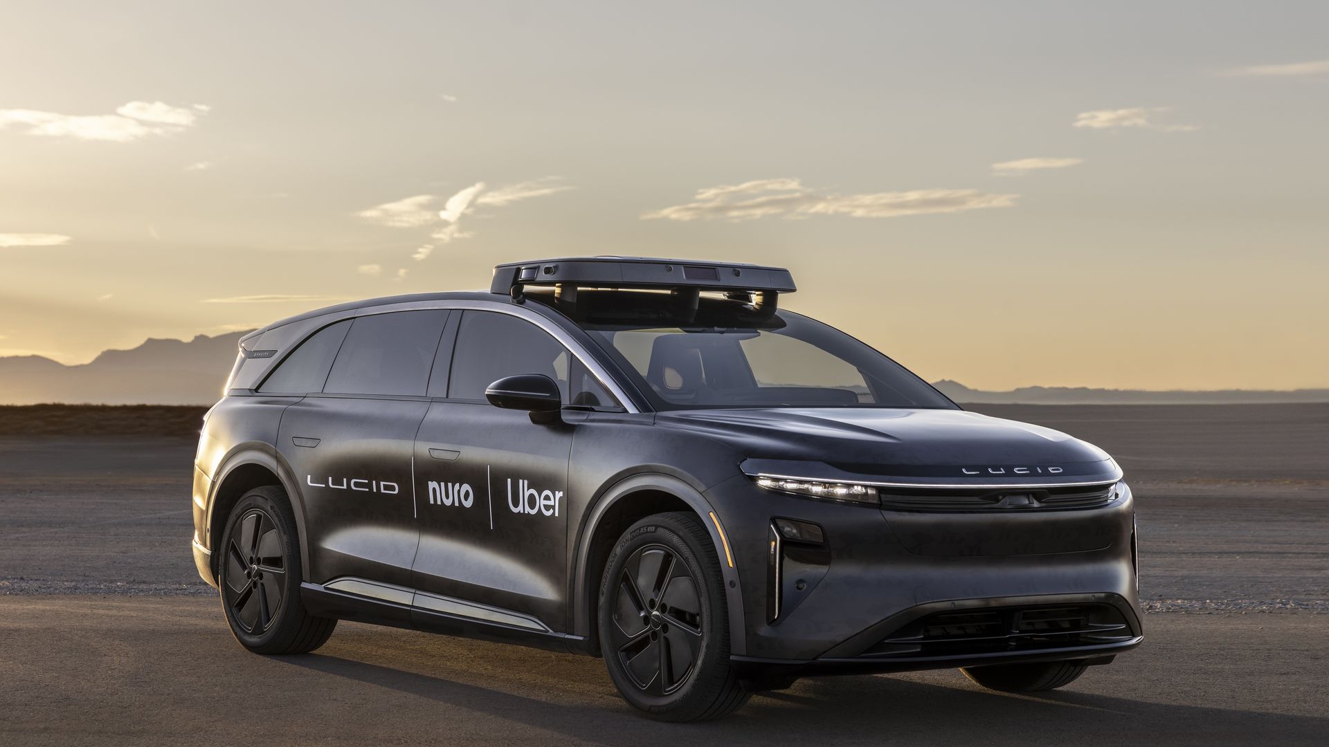 Black Lucid electric SUV with Nuro and Uber logos, equipped with rooftop autonomous driving sensors, parked on a desert road at sunset with mountains in the background.