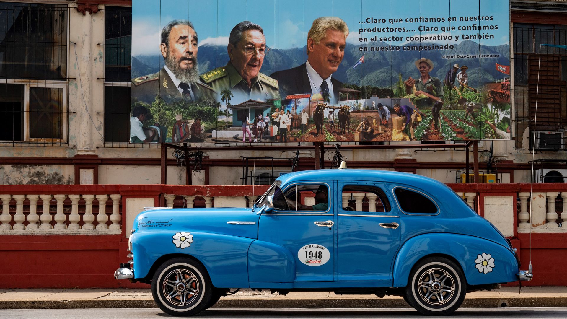 Bright blue vintage car with white daisy decals sits in front of a large mural featuring three men in uniforms ( late Cuban leader Fidel Castro (L), former President Raul Castro (C) and current President Miguel Diaz-Canel ), village scenes, and a mountains backdrop.