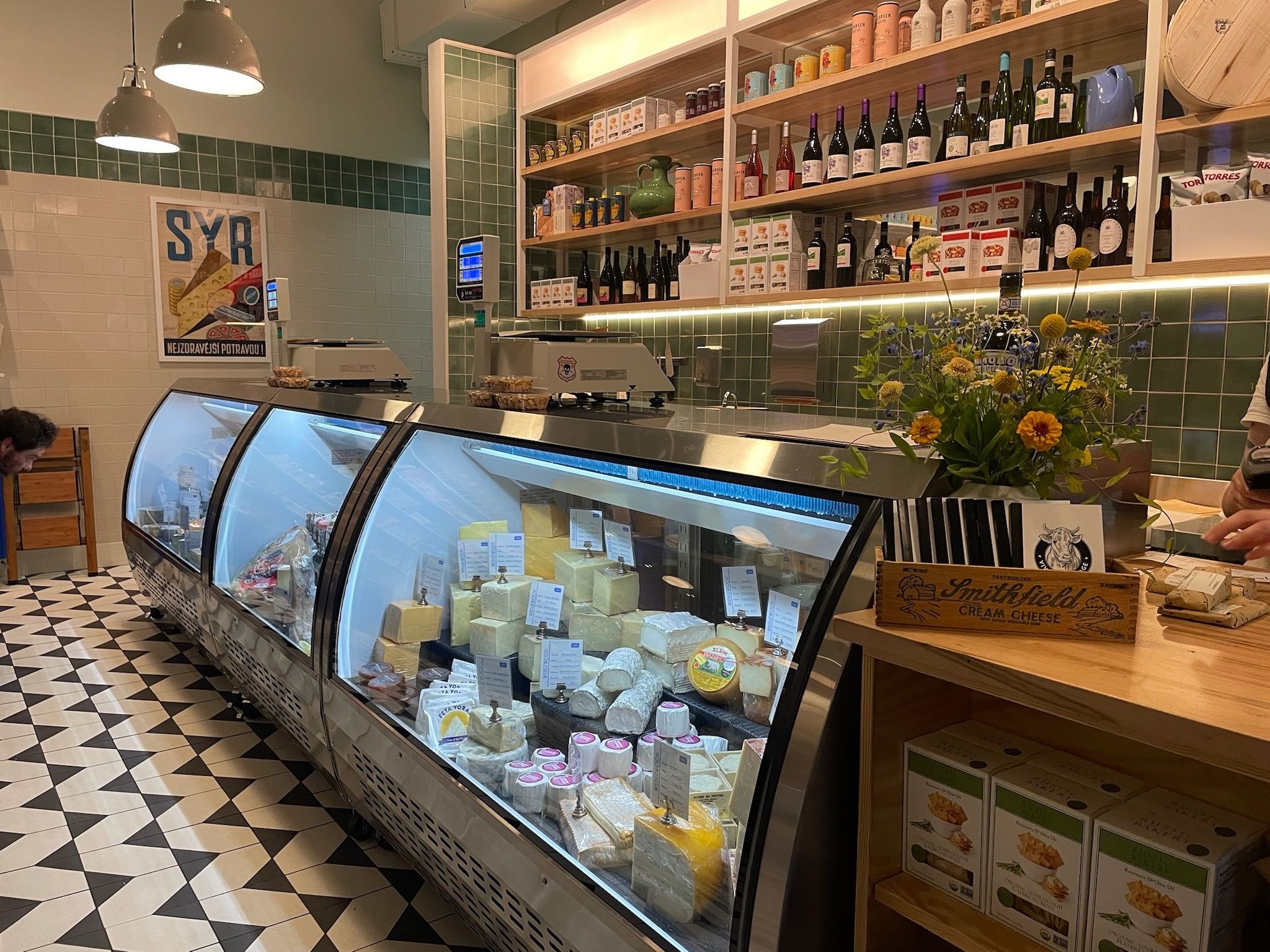 Interior of a cheese shop with a glass display case filled with various cheeses, green tiled walls, wooden shelves with wines and products, and a flower arrangement on the counter.