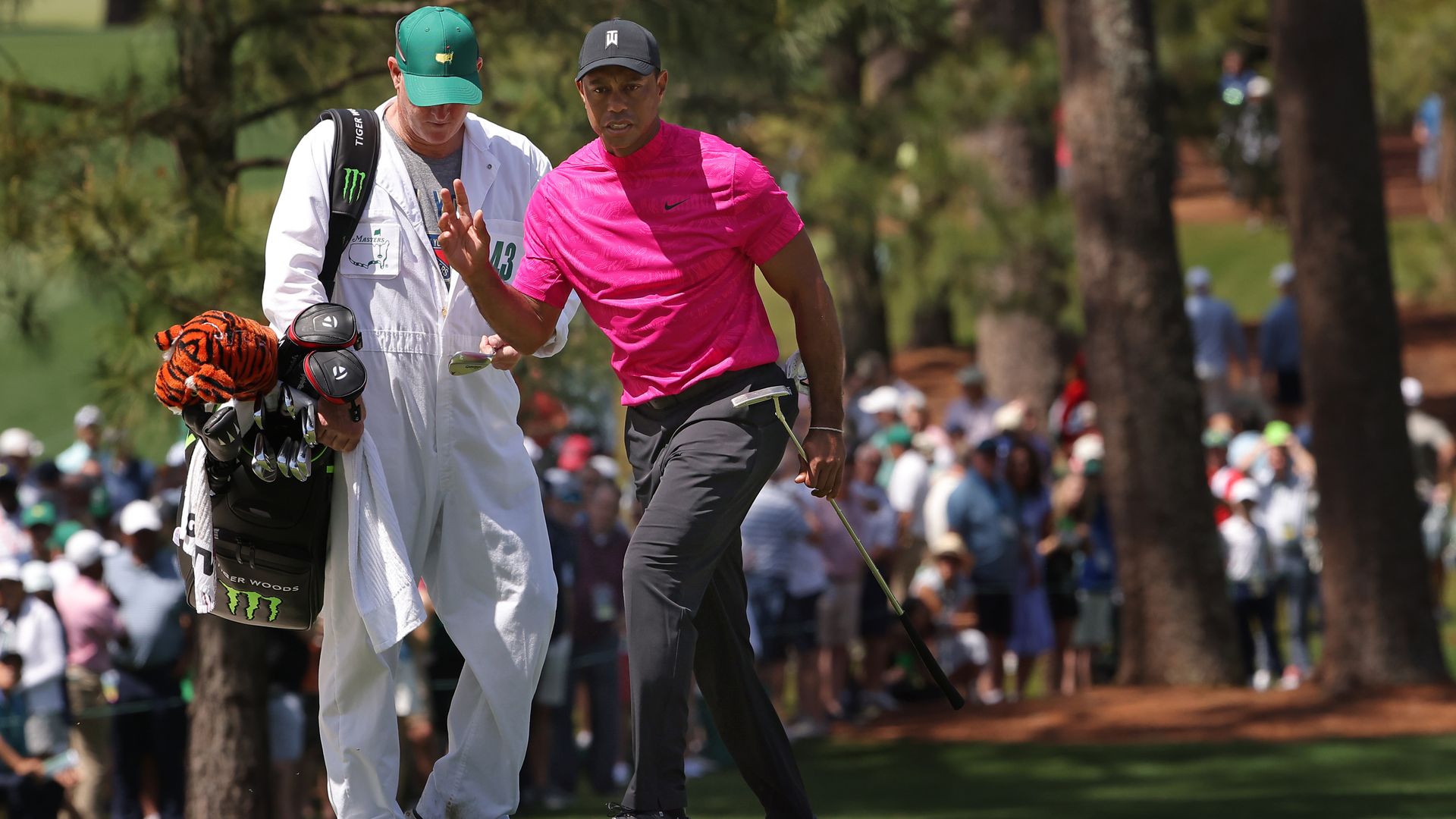 A golfer in a pink shirt waves to the crowd with his caddie, in a white jumpsuit, behind him