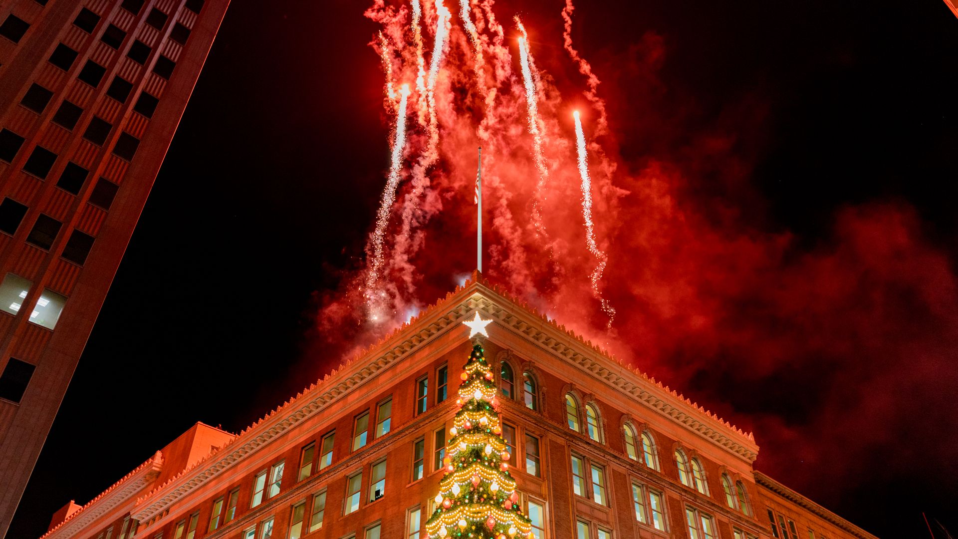 Nighttime scene of fireworks bursting in red above a decorated Christmas tree topped with a star in front of a large brick building with lit windows.
