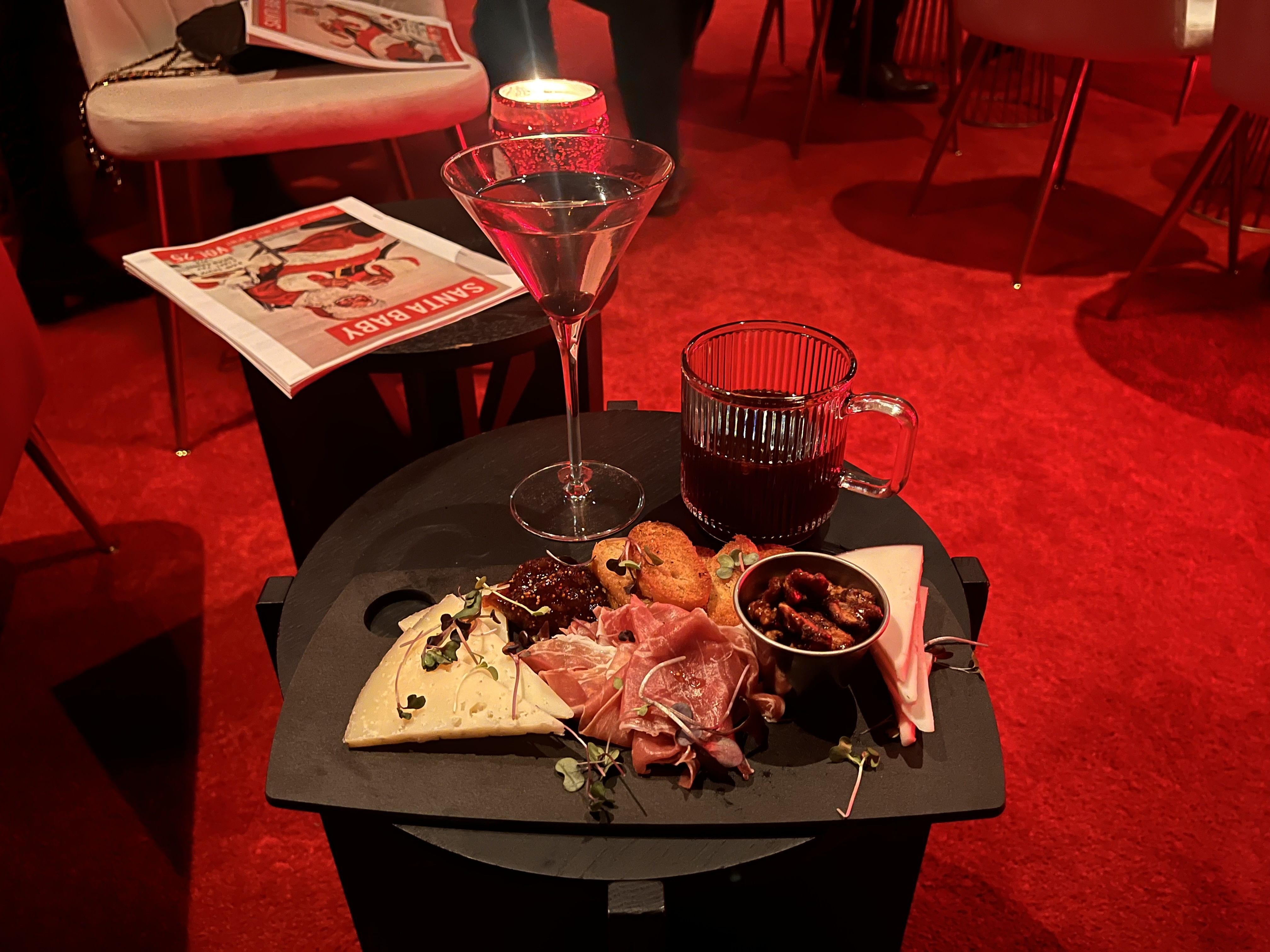 Charcuterie board with cheese, prosciutto, nuts, crackers, and jam on a black table with a martini glass and a coffee mug, under warm red lighting.