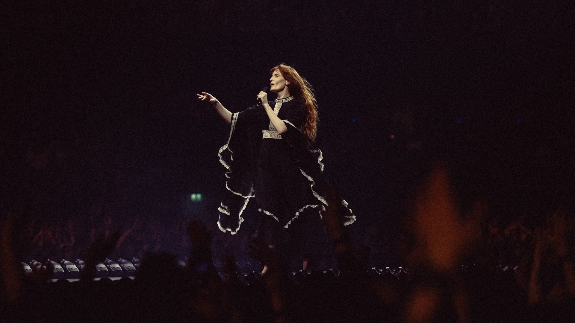Florence Welch of 'Florence And The Machine' in a black robe with white trim, holding a microphone on a dimly lit stage, performing as a dark audience sits in the foreground.