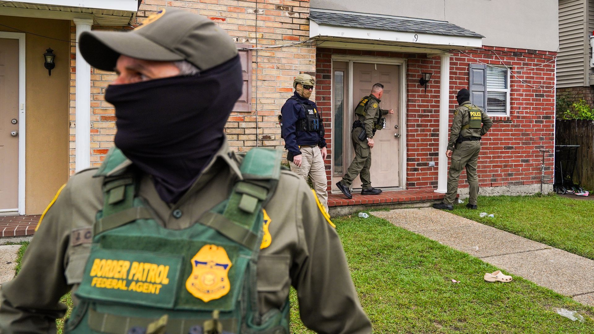 Three federal agents approach a residential door in the background as a fourth agent, with his face mostly covered, stands in the foreground.