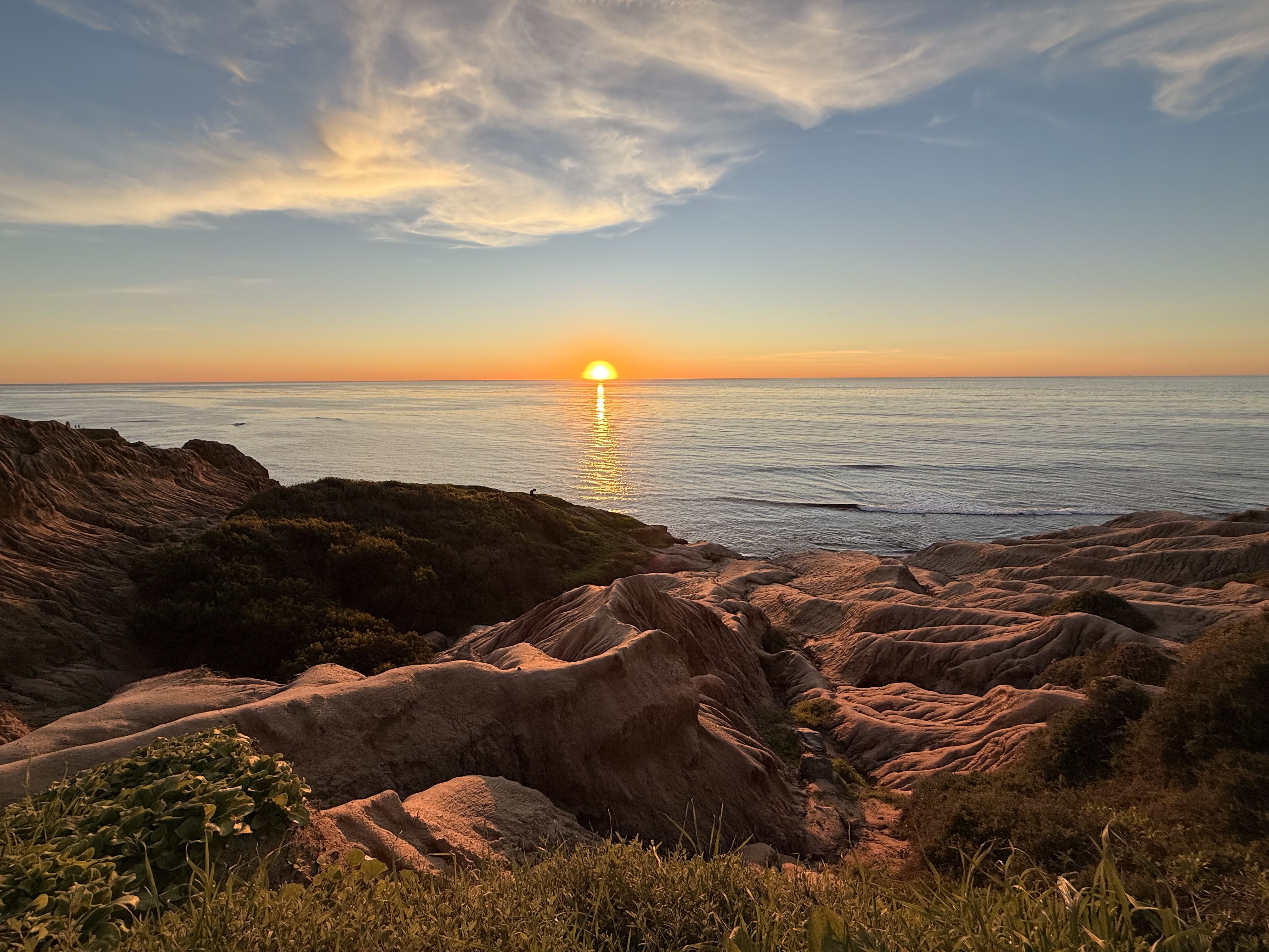 Golden sun setting over tranquil sea, reflecting orange light on water. Jagged rocky coastline with green shrubs in foreground under a pale blue sky with wispy clouds.