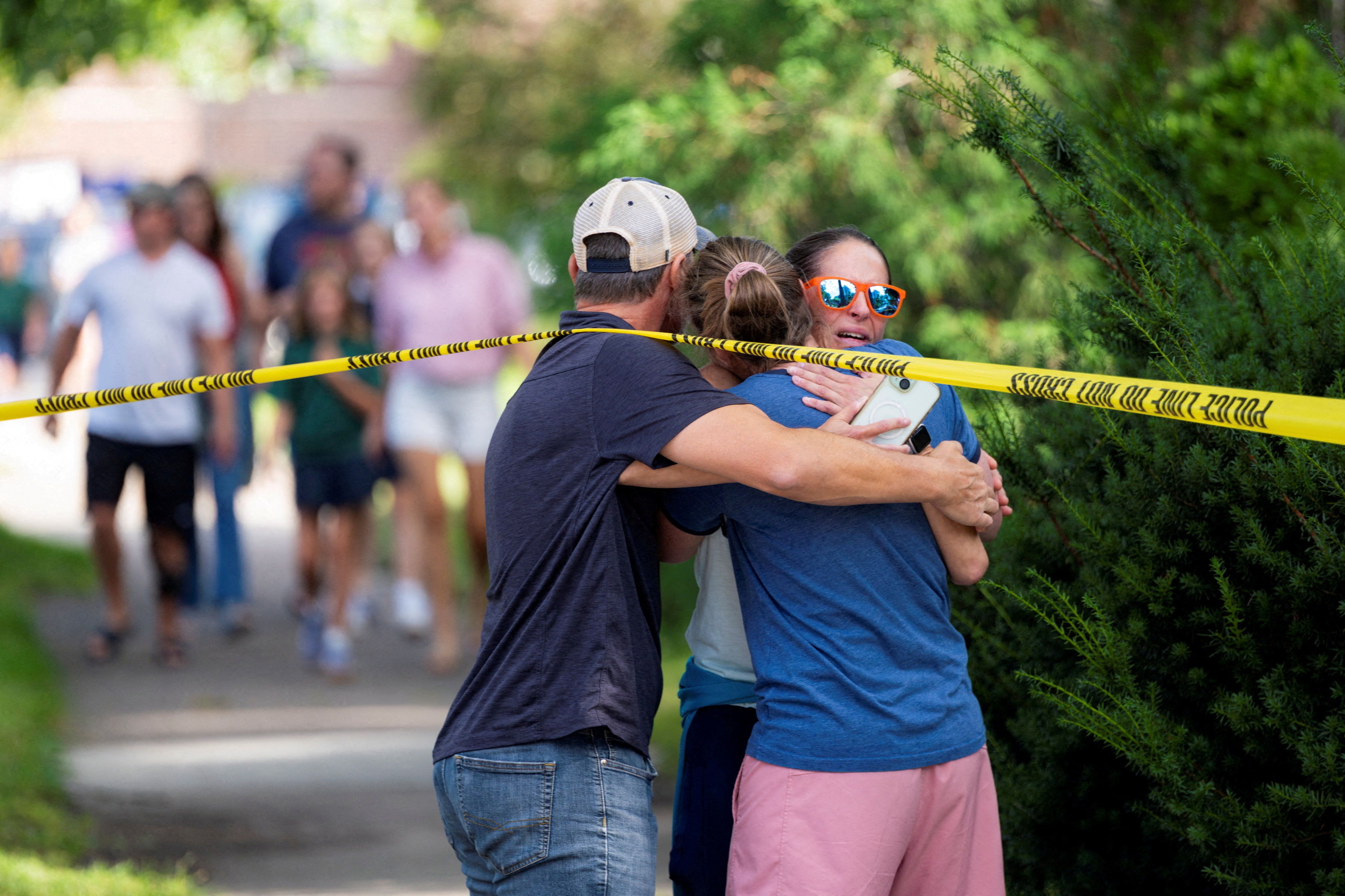 Three people hug in front of yellow police tape with blurred people in the background on a sunny day with green trees around.