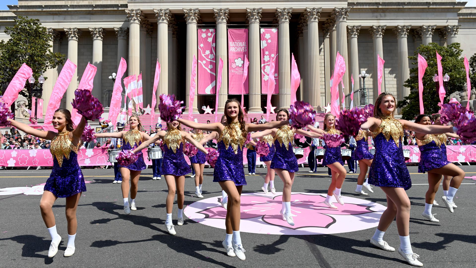Cheerleaders in purple and gold sequined dresses perform dancing routine with pink pom-poms and flags outside the National Archives building during the National Cherry Blossom festival.
