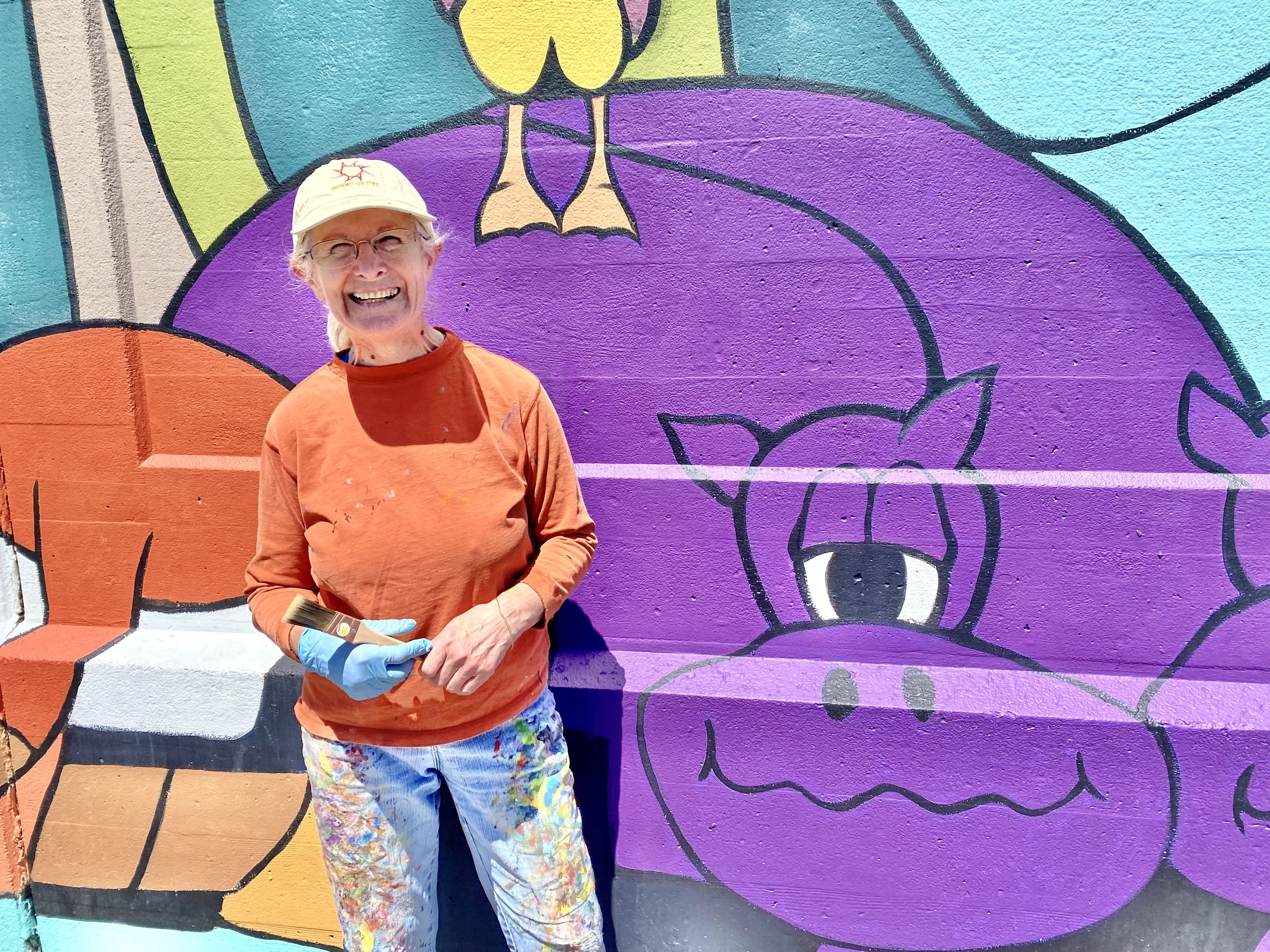 A woman in an orange-red shirt and paint stained pants stands next to two purple hippos in a mural.