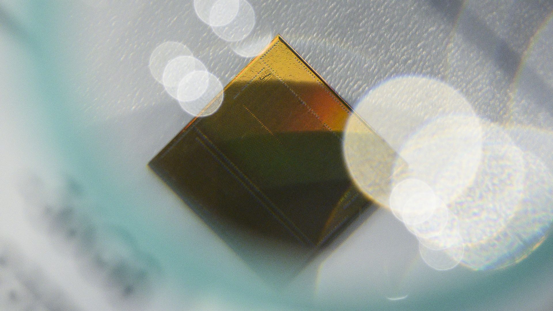 Macro shot of a gold, diamond-shaped metal edge with fine etched lines, set against a soft teal-blue background and bright circular bokeh lights.