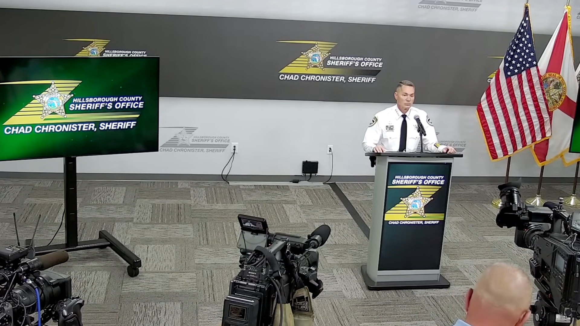 Sheriff in white uniform at podium during Hillsborough County Sheriff's Office press conference with American and Florida flags, cameras recording, and office logos visible.