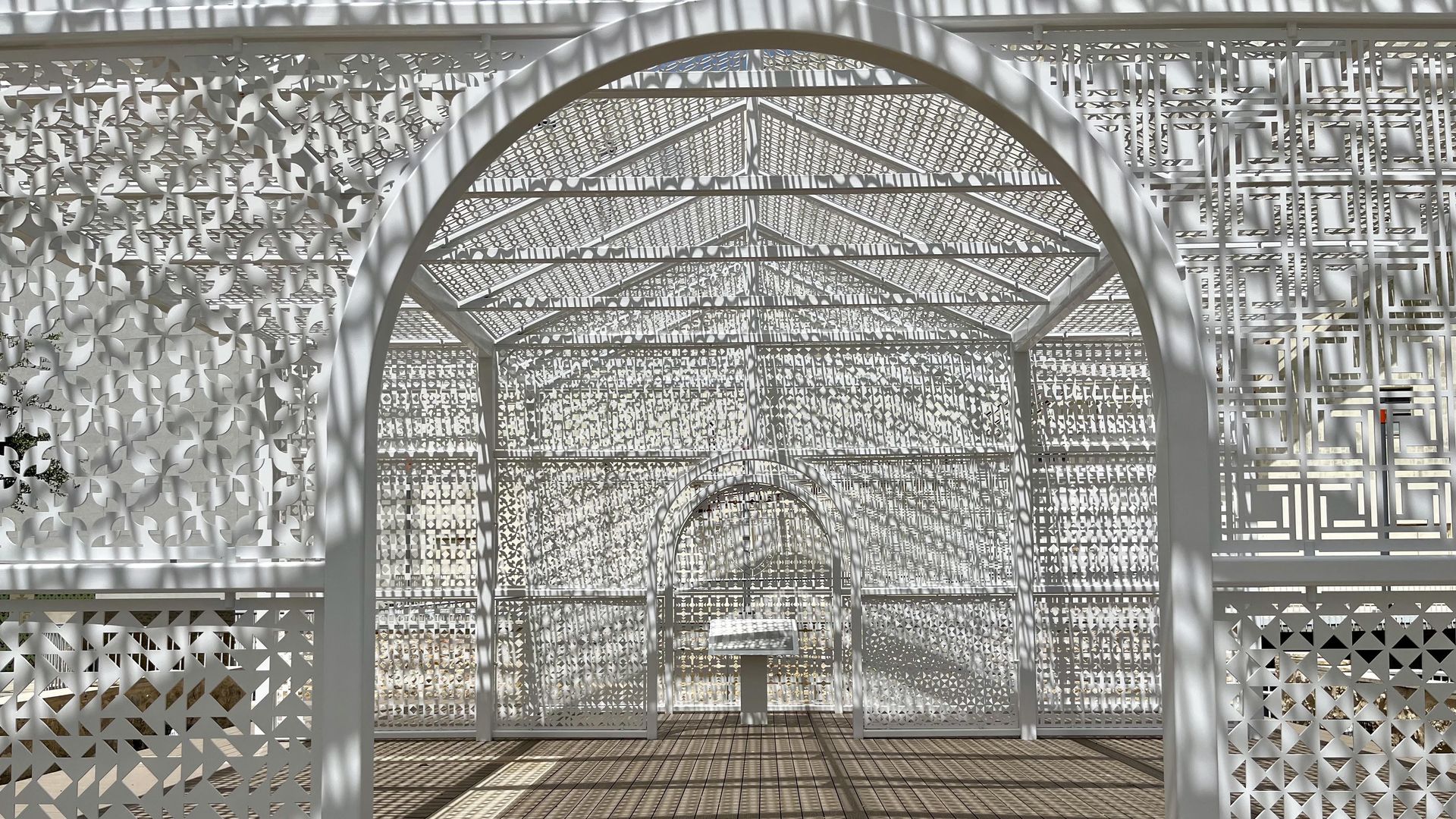 A sunlit white lattice walkway with arches and intricate geometric quilt panels. Light filters through perforations, casting striped shadows on the wooden floor as the tunnel stretches inward. "Presence of the Past" by artist Gordon Huether on the San Pedro Creek honors an early Black church.