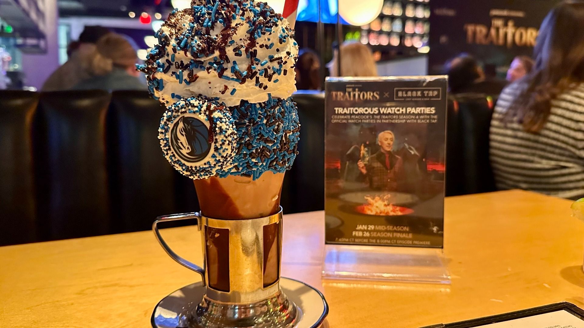 Tall glass milkshake with whipped cream, blue and brown sprinkles, and a cookie with a sports team logo, on a table at a restaurant with a "Traitors" watch party sign in the background.