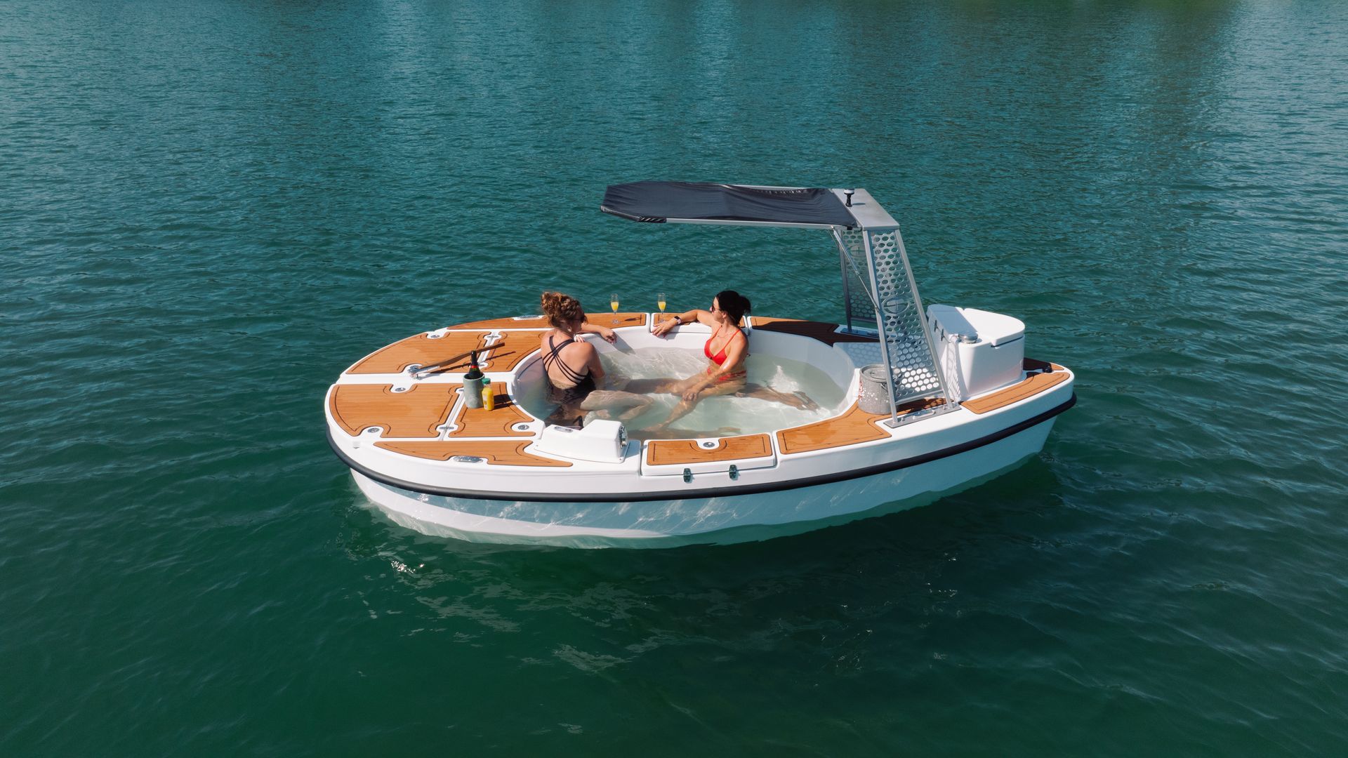Two women in swimsuits, one in a red bikini and the other in a black striped suit, relax in a round hot tub on a wooden-deck yacht, sipping drinks as turquoise water surrounds them.