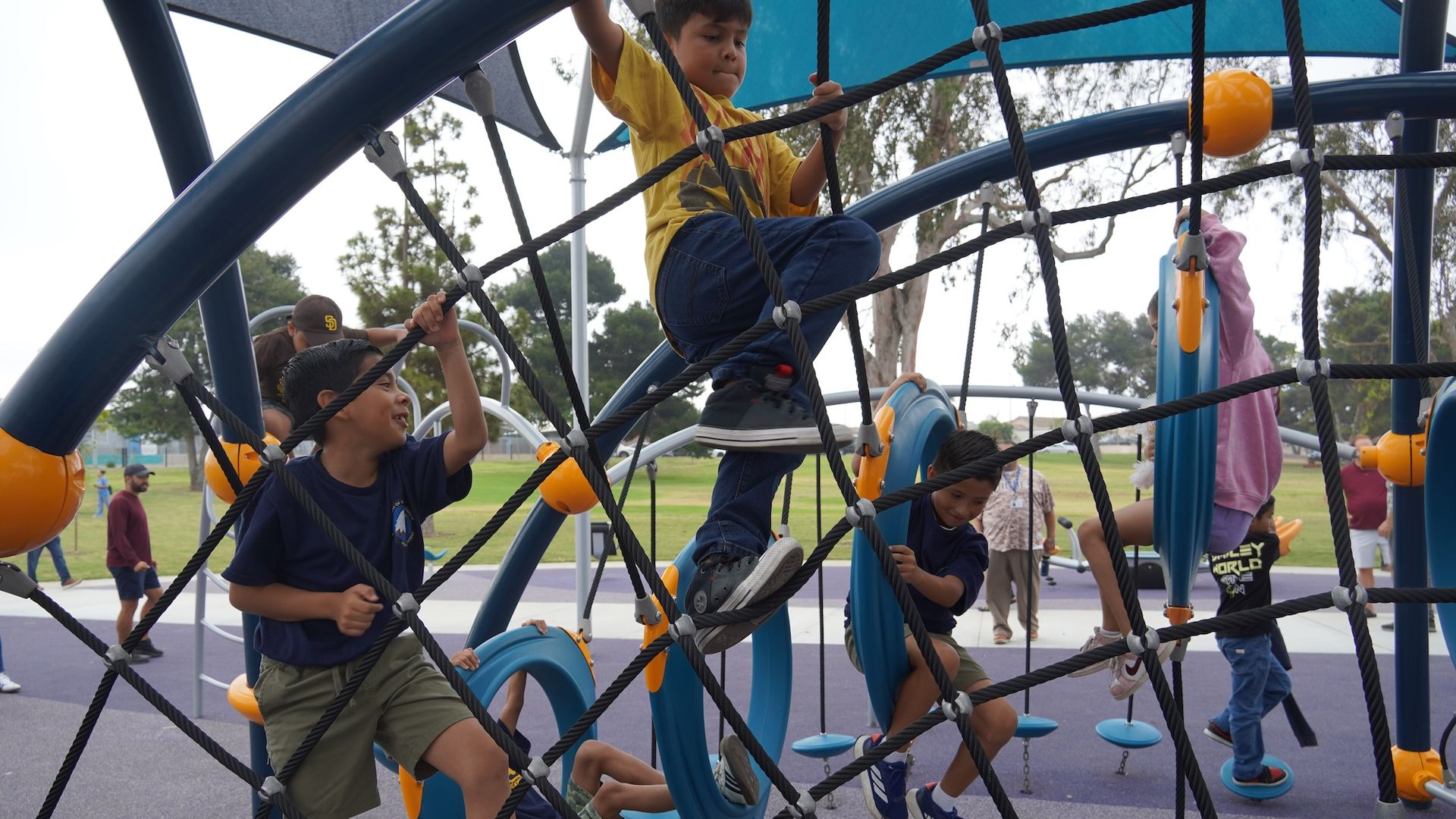 Children climbing and playing on blue and orange playground equipment with rope nets and circular seats, outdoors in a park with green grass and trees under a blue canopy.