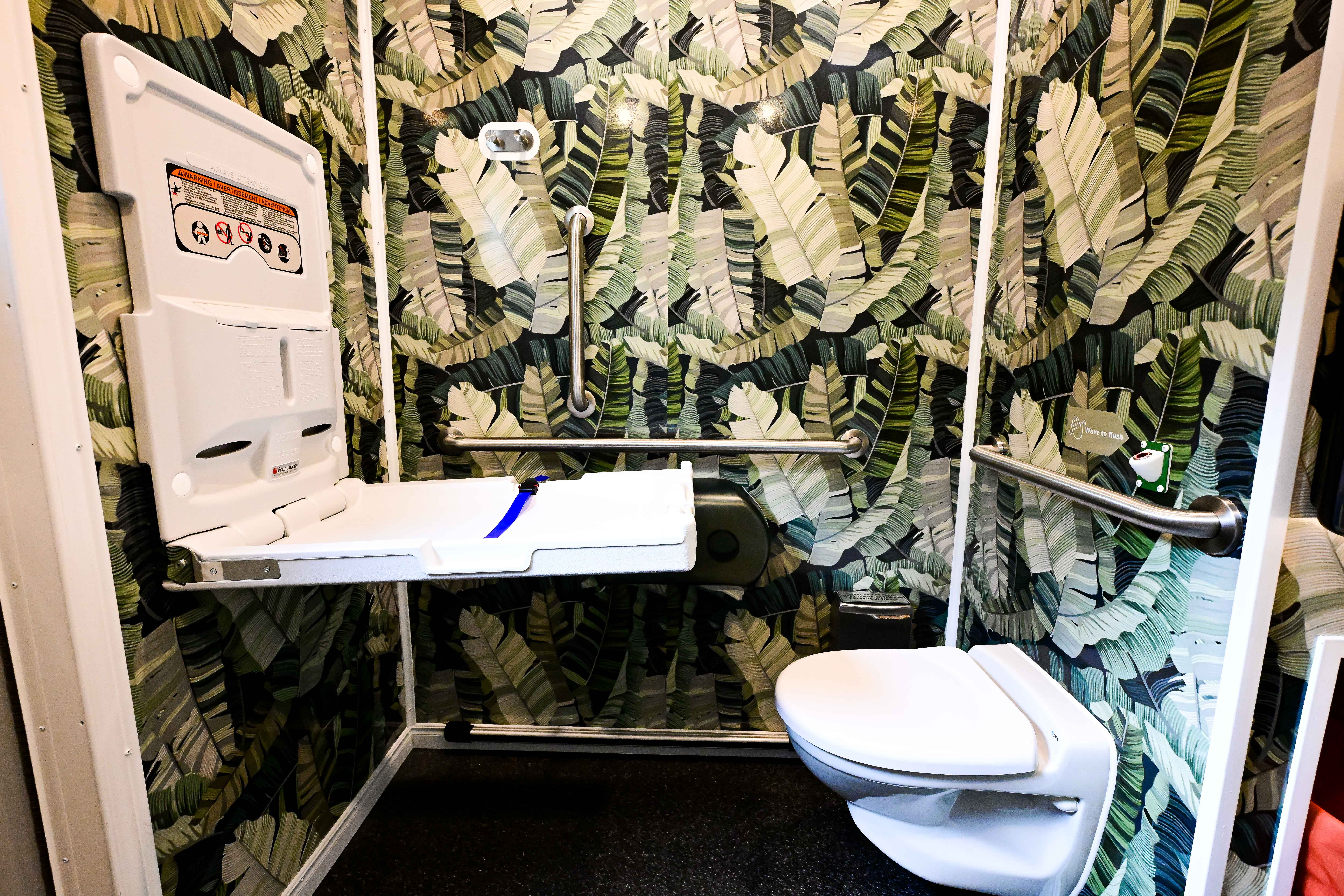 A compact lavatory with bold tropical leaf wallpaper, a fold-down baby changing table on the left, chrome grab bars, and a white toilet on the right.