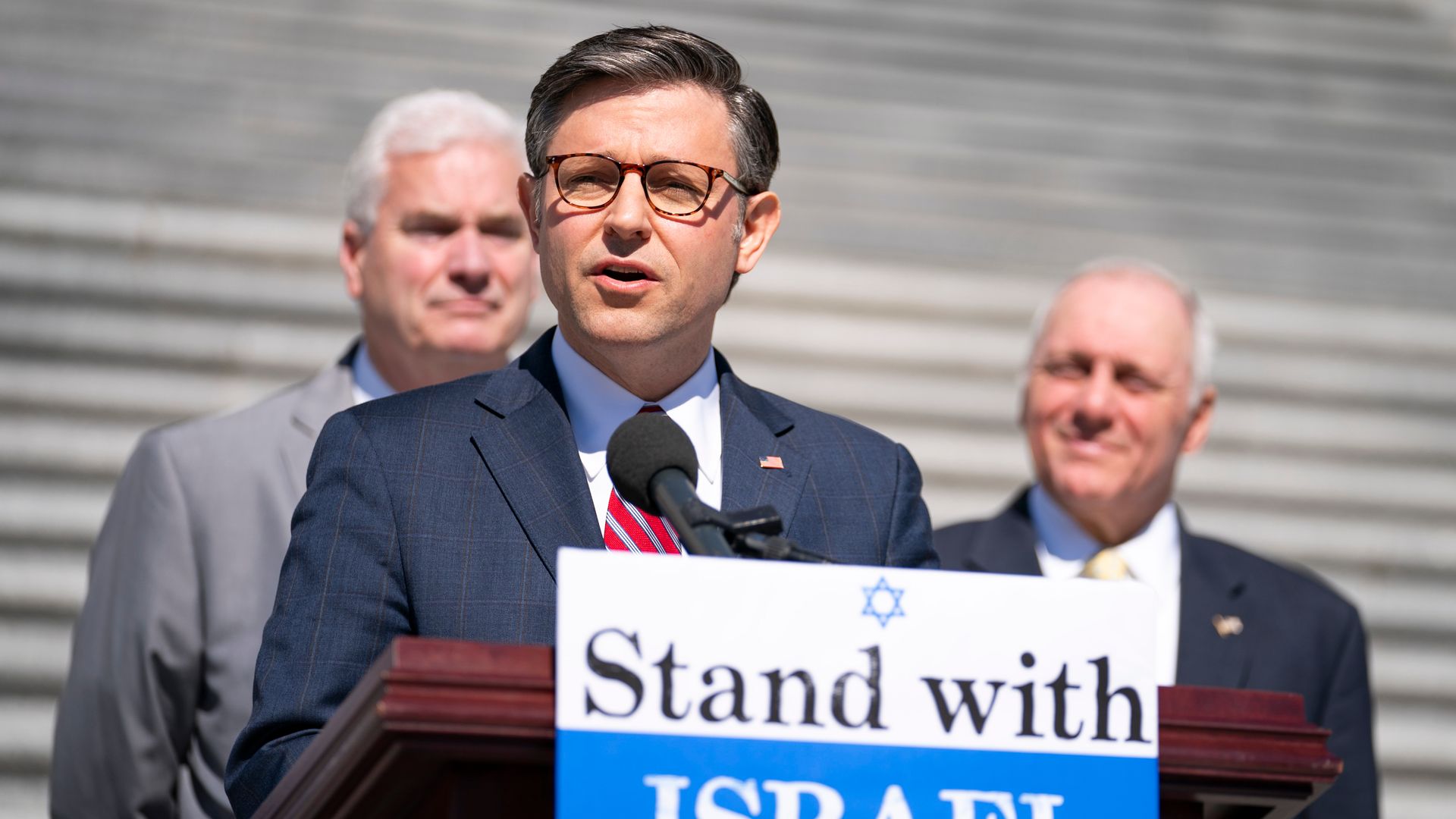 Speaker Mike Johnson, wearing a blue suit, white shirt and red striped tie, speaking at a podium with a "stand with Israel" poster on it flanked by colleagues in front of stairs.