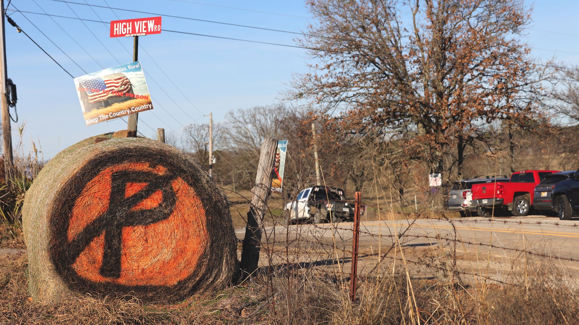 Round hay bale painted with orange background and black symbol crossed by a line near High View Road sign, rural road with parked trucks and leafless trees under clear sky.