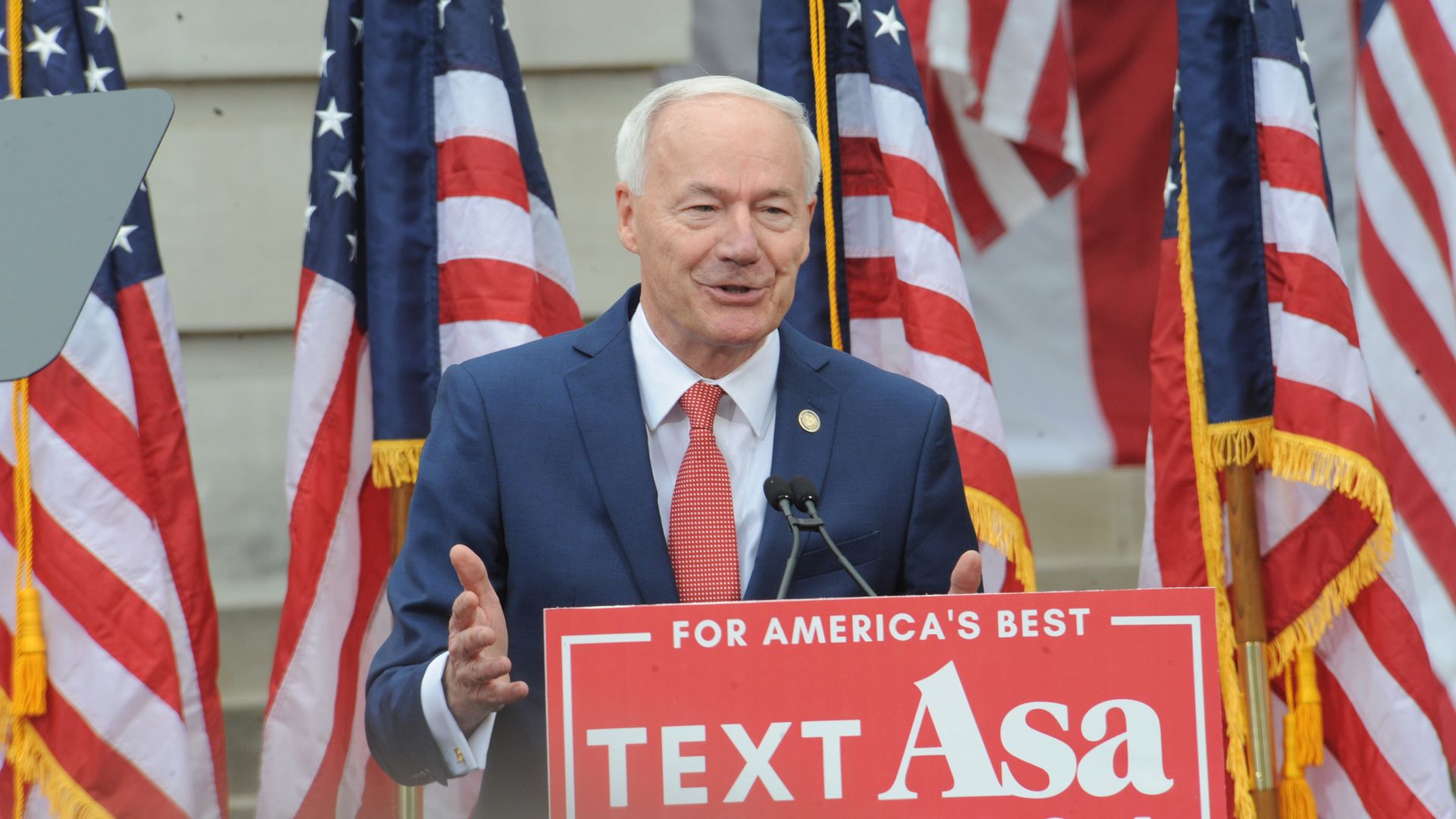 A photo of former Arkansas Gov. Asa Hutchinson speaking at a podium surrounded by American flags.