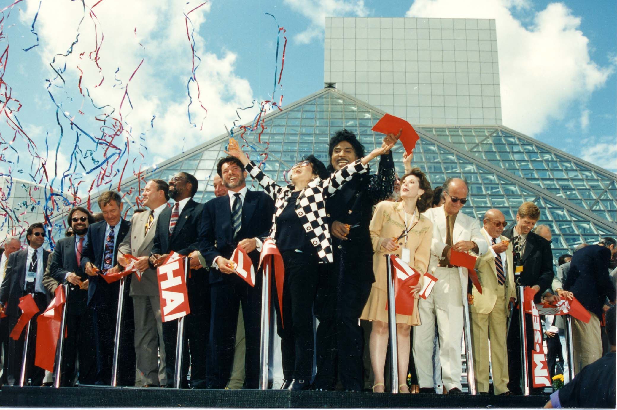 Group of professionals on a platform outside a glass pyramid, celebrating with red ribbons and streamers; central figure in a black-and-white checkered jacket raises arms in triumph.