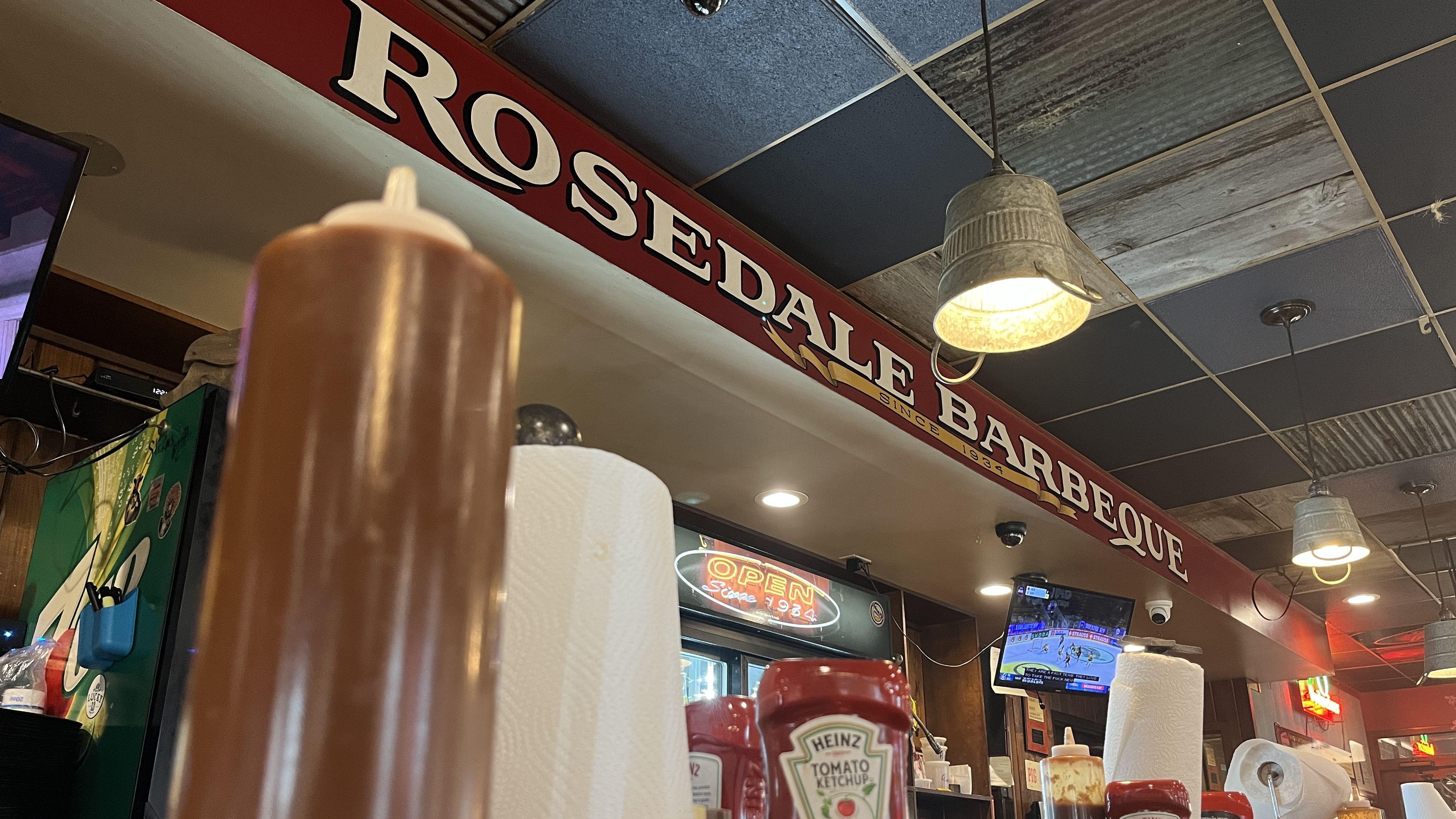 Inside Rosedale Barbeque with red sign, metal hanging lamps, condiments including ketchup, barbecue sauce, and paper towels on tables, and a TV showing a sports game.