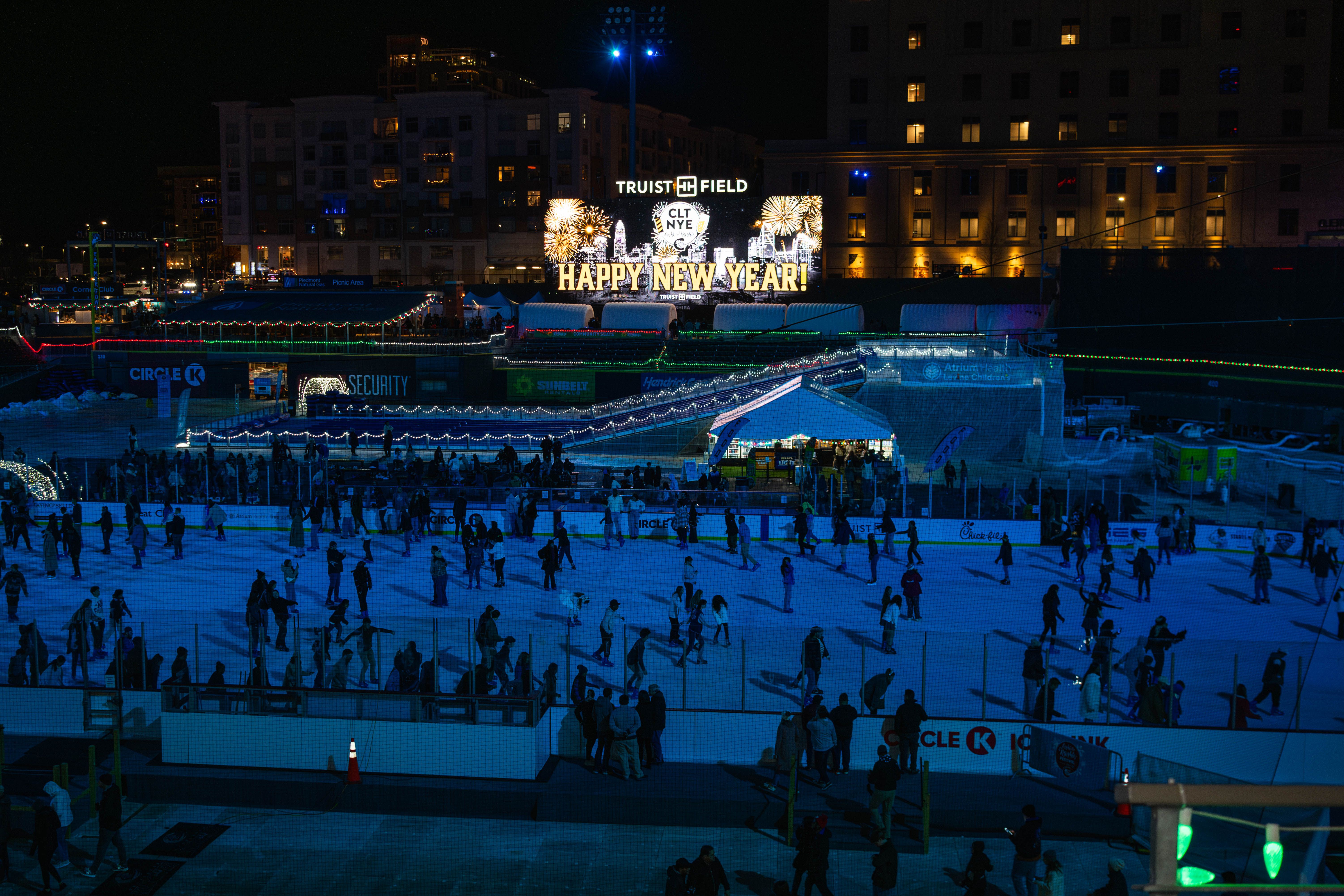 People ice skating at night on a rink lit blue at Truist Field with a lit sign reading "Happy New Year!" and fireworks graphics in the background, surrounded by buildings and lights.