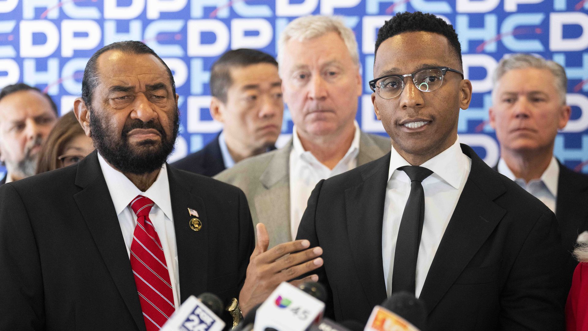 Group of men in suits at a press conference; foreground left man wears a red striped tie, right man wears glasses; microphones in front; blue backdrop with white letters.