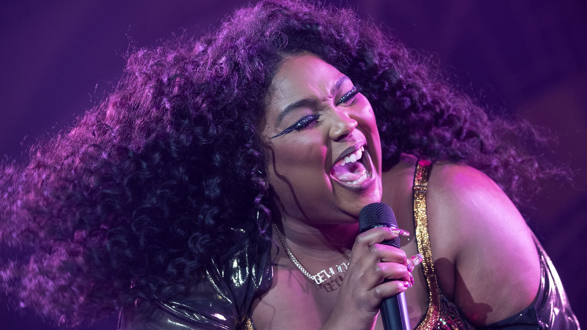 Lizzo sings into a microphone with purple light shining on her face.