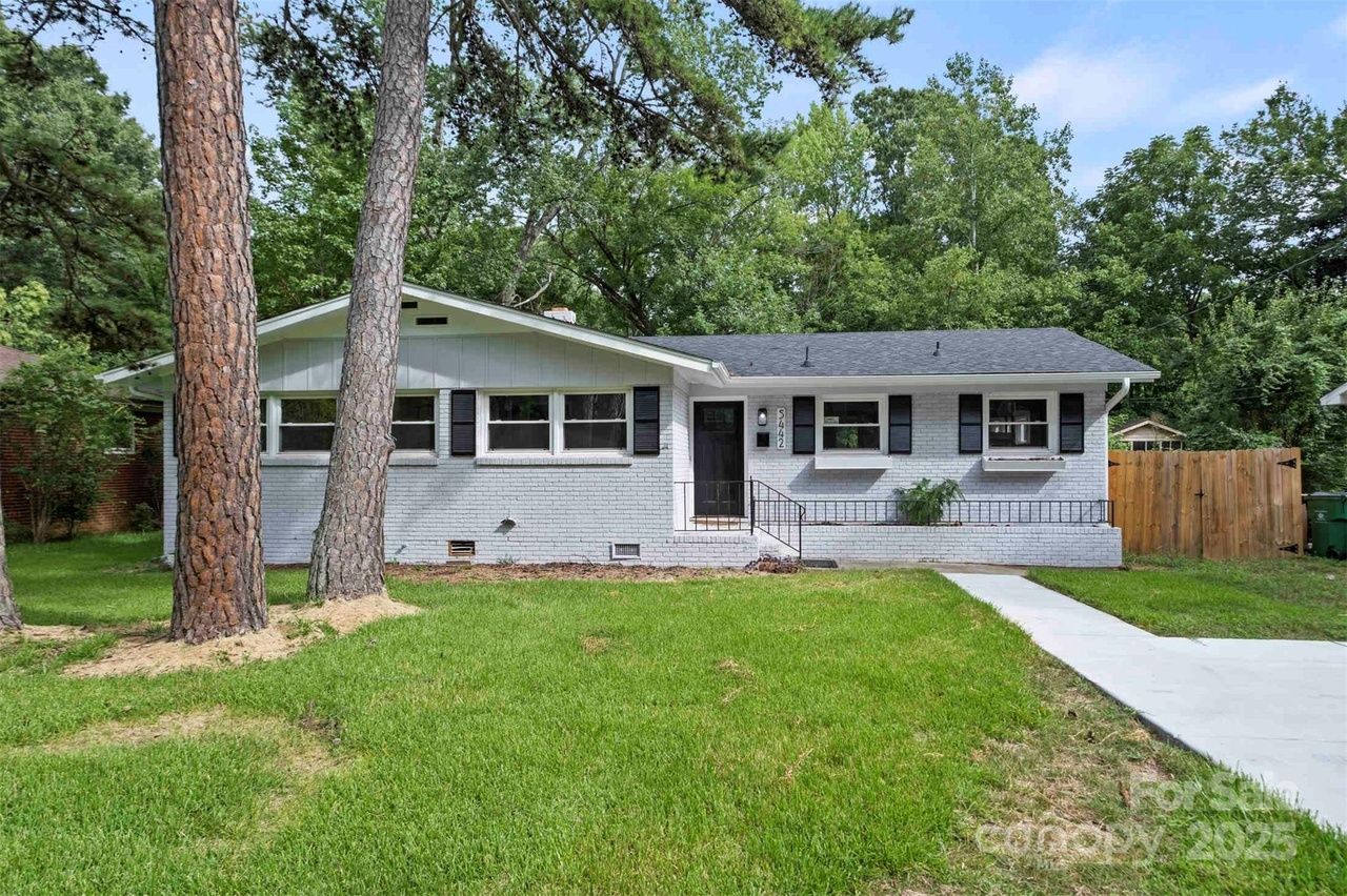 Single-story white brick house with black shutters, a black front door, and a gray roof, set in a green lawn with tall pine trees and a wooden fence in the background.