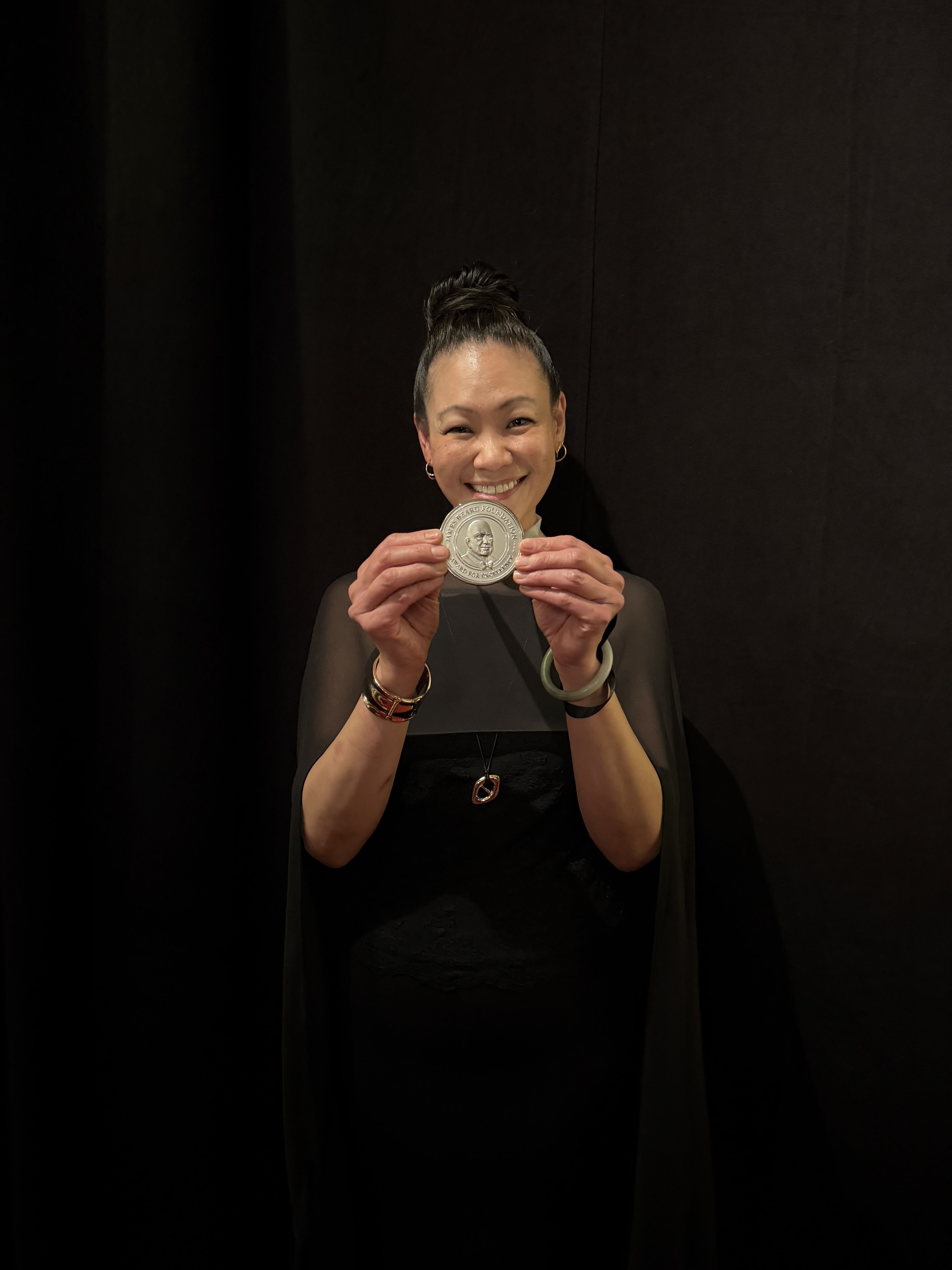 Smiling woman with her hair in a bun holds a large silver medal toward the camera. She wears hoop earrings and bracelets, in a black outfit, against a dark stage backdrop.