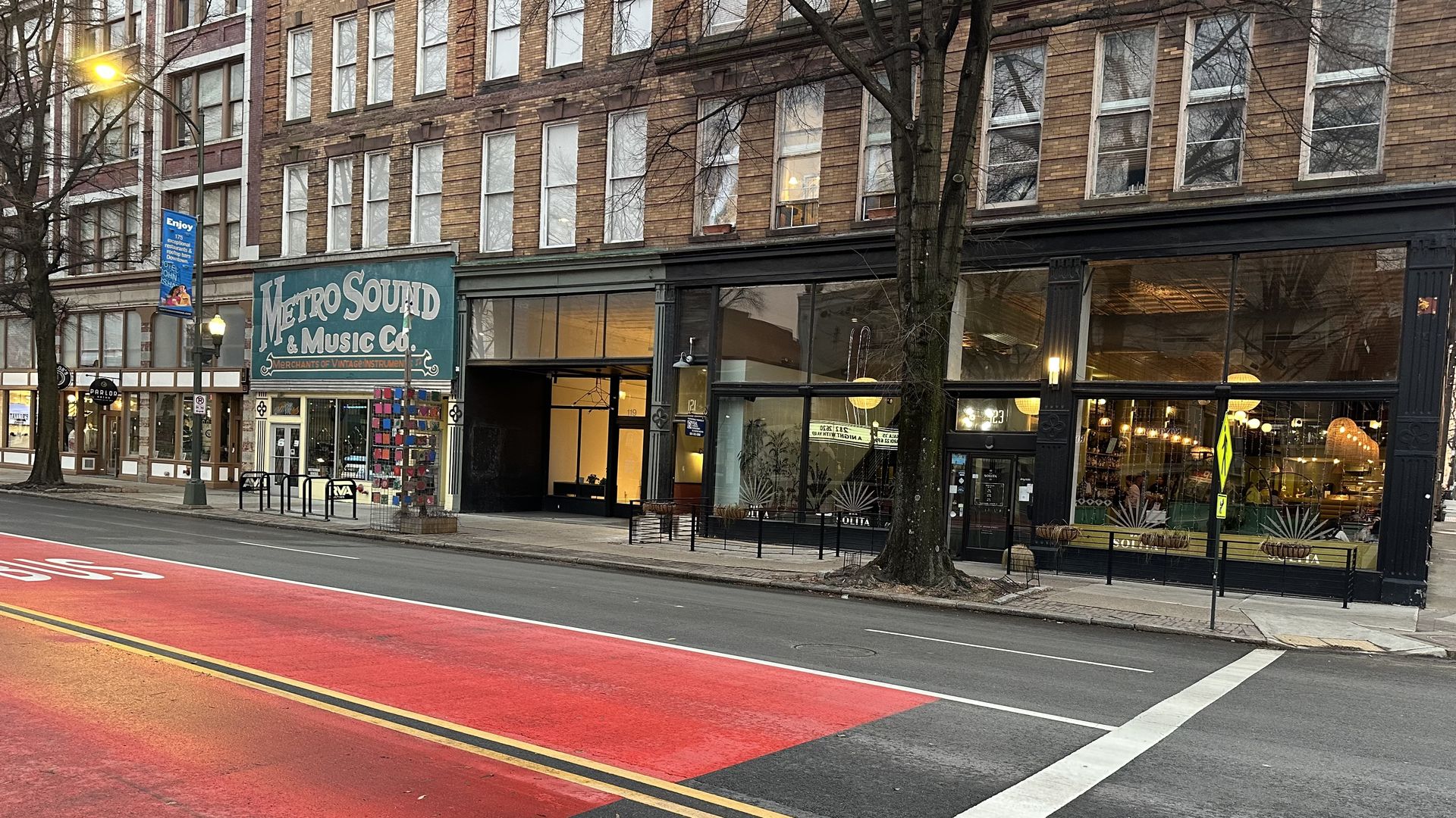 City street with a brick storefront building and large glass windows. A teal sign reads "Metro Sound & Music Co." under the store; a blue banner hangs from a streetlight. Bare trees line the sidewalk; a red bus lane runs by.