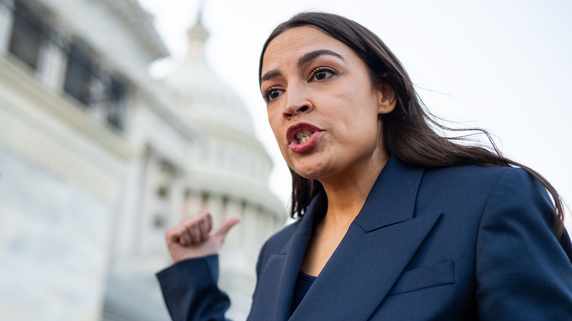 A woman with long dark hair wearing a navy blazer speaks outdoors, her mouth open in mid-sentence, gesturing with a raised hand as a stone building blurs in the background.