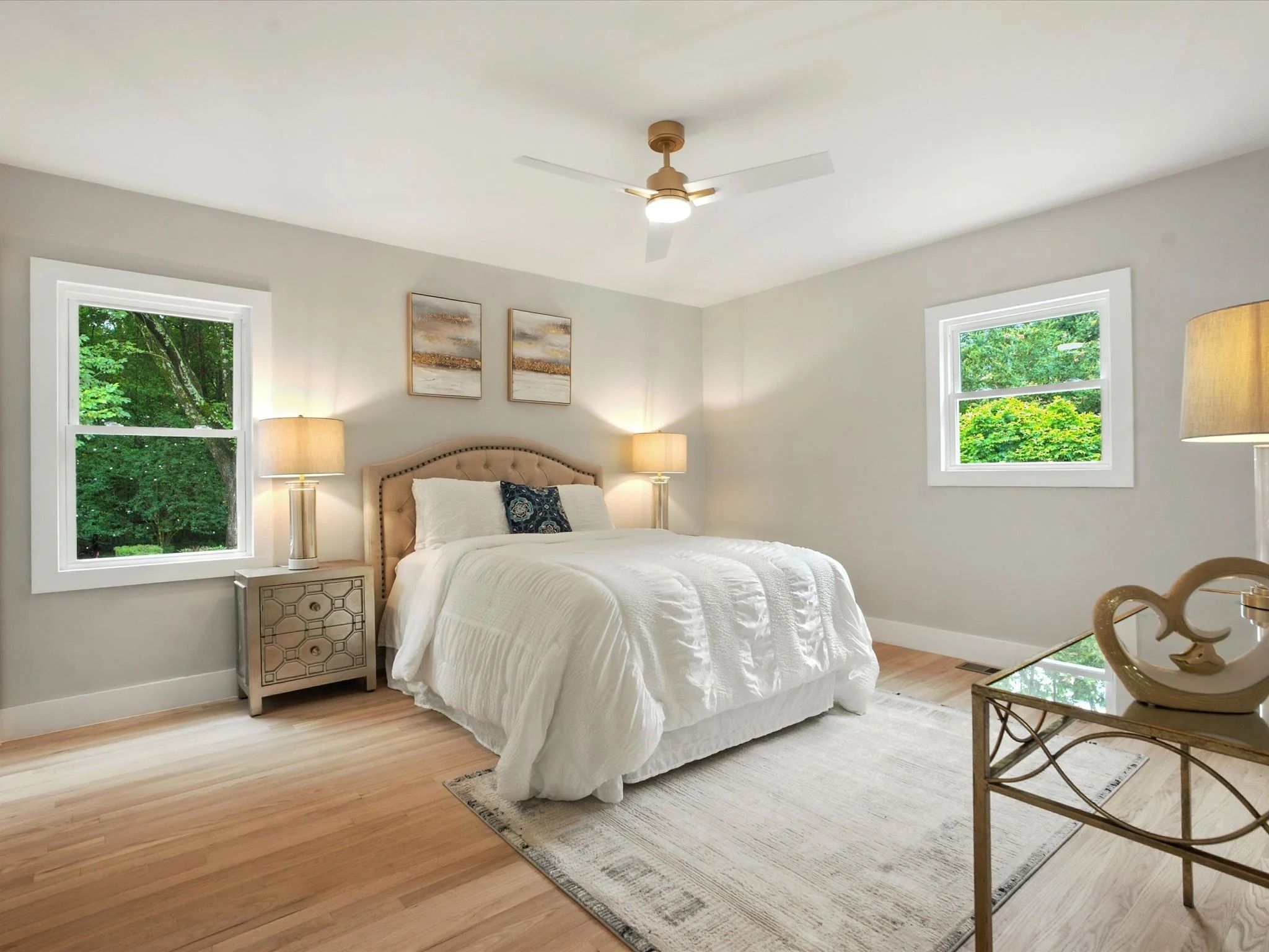 Bright bedroom with white bedding, beige tufted headboard, two lamps on nightstands, two windows showing green trees, light wood floor, abstract art, and ceiling fan with light.