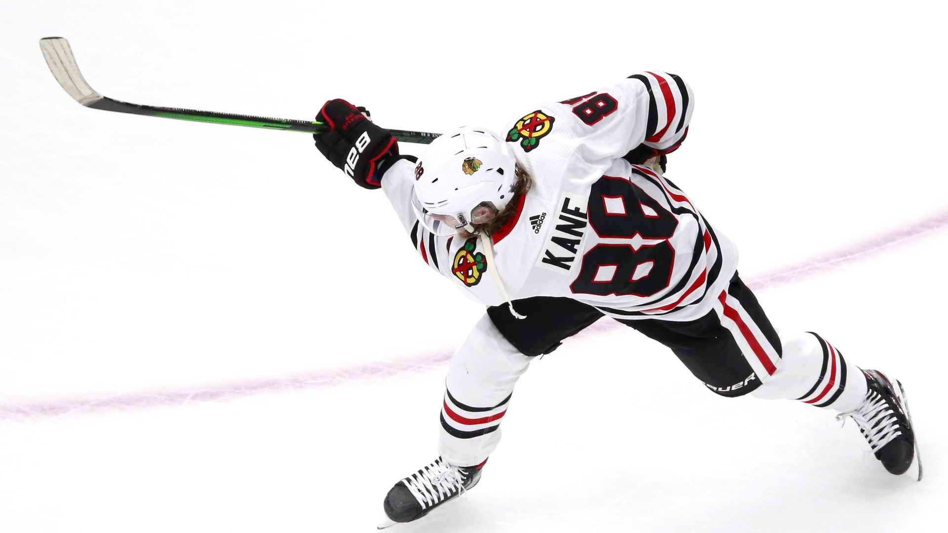 Hockey player in a white Chicago Blackhawks jersey number 88 with Kane on the back, wearing black gloves and skating while gripping a green hockey stick on the ice.