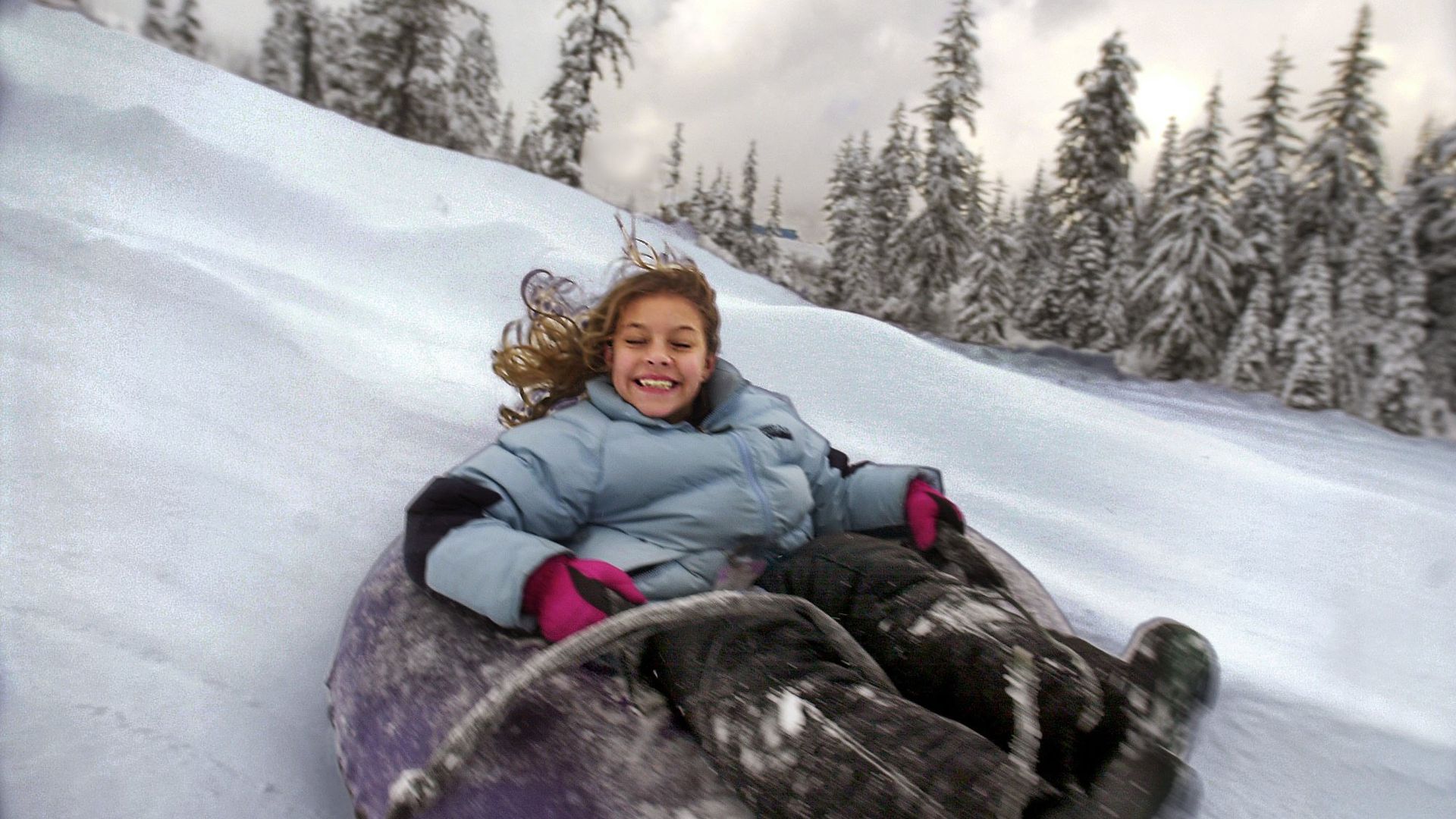 A child rides down a snowy hill in an innertube while laughing and smiling. 