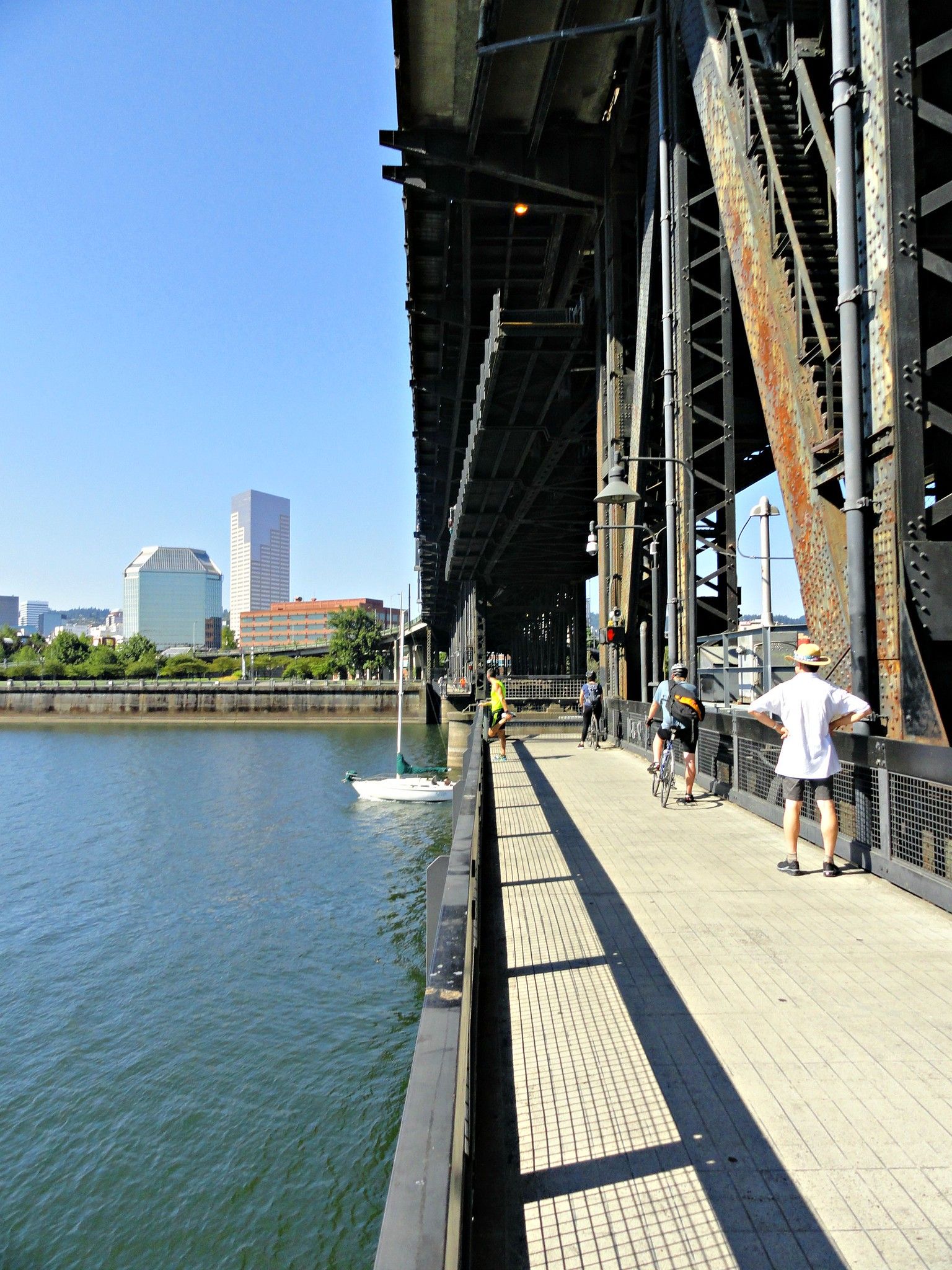Rusted steel bridge looms over a riverside pedestrian and bike path; people walk and cycle as a distant city skyline and blue sky appear.