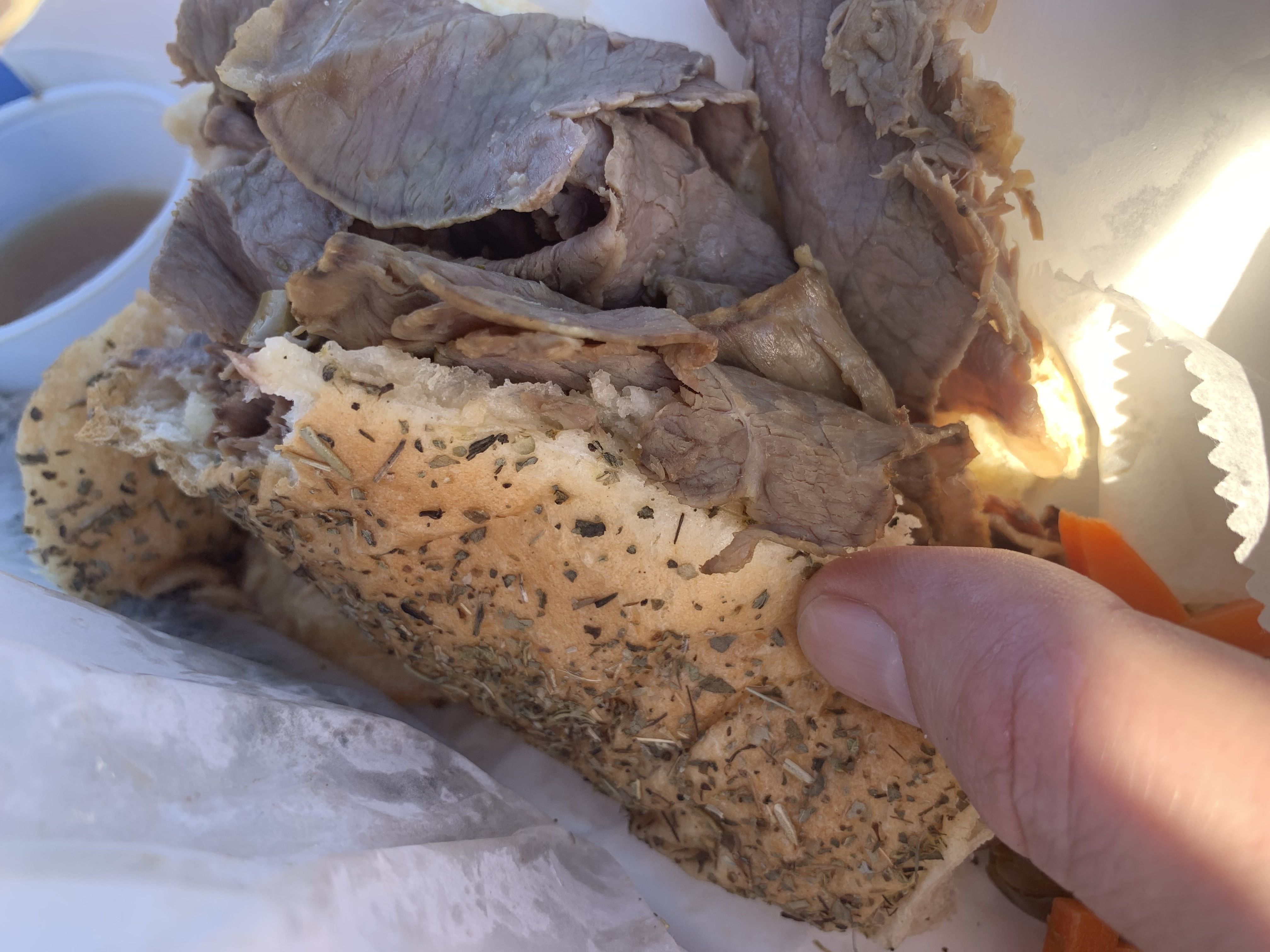 Hand holding an Italian beef sandwich with seasoned bread, thinly sliced roast beef, and a side of au jus and pickled carrots in white containers.