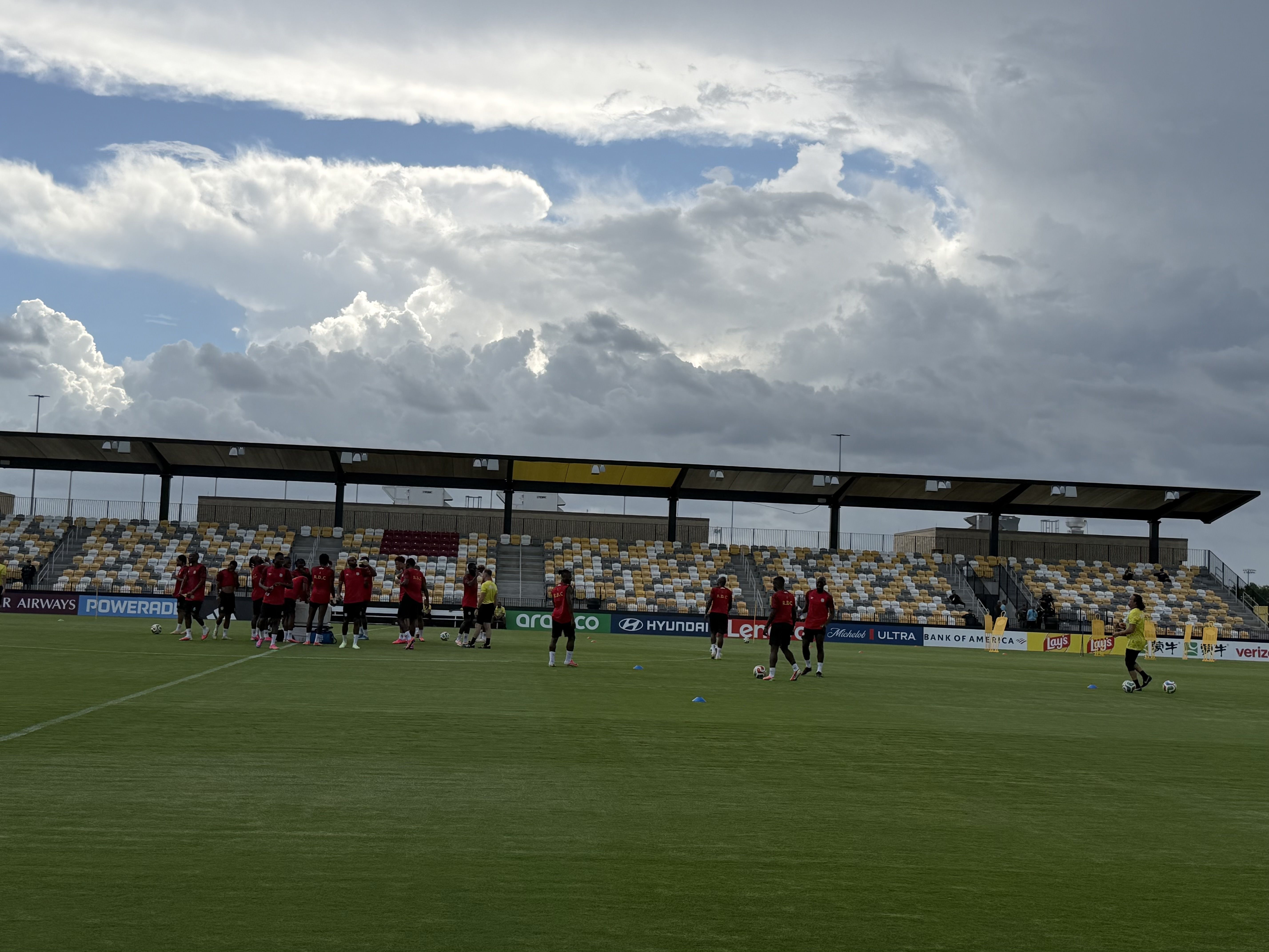 Soccer stadium with players in red jerseys gathered on a green field, sidelines lined with advertising boards; yellow and white seats, covered stands, and a cloudy sky.